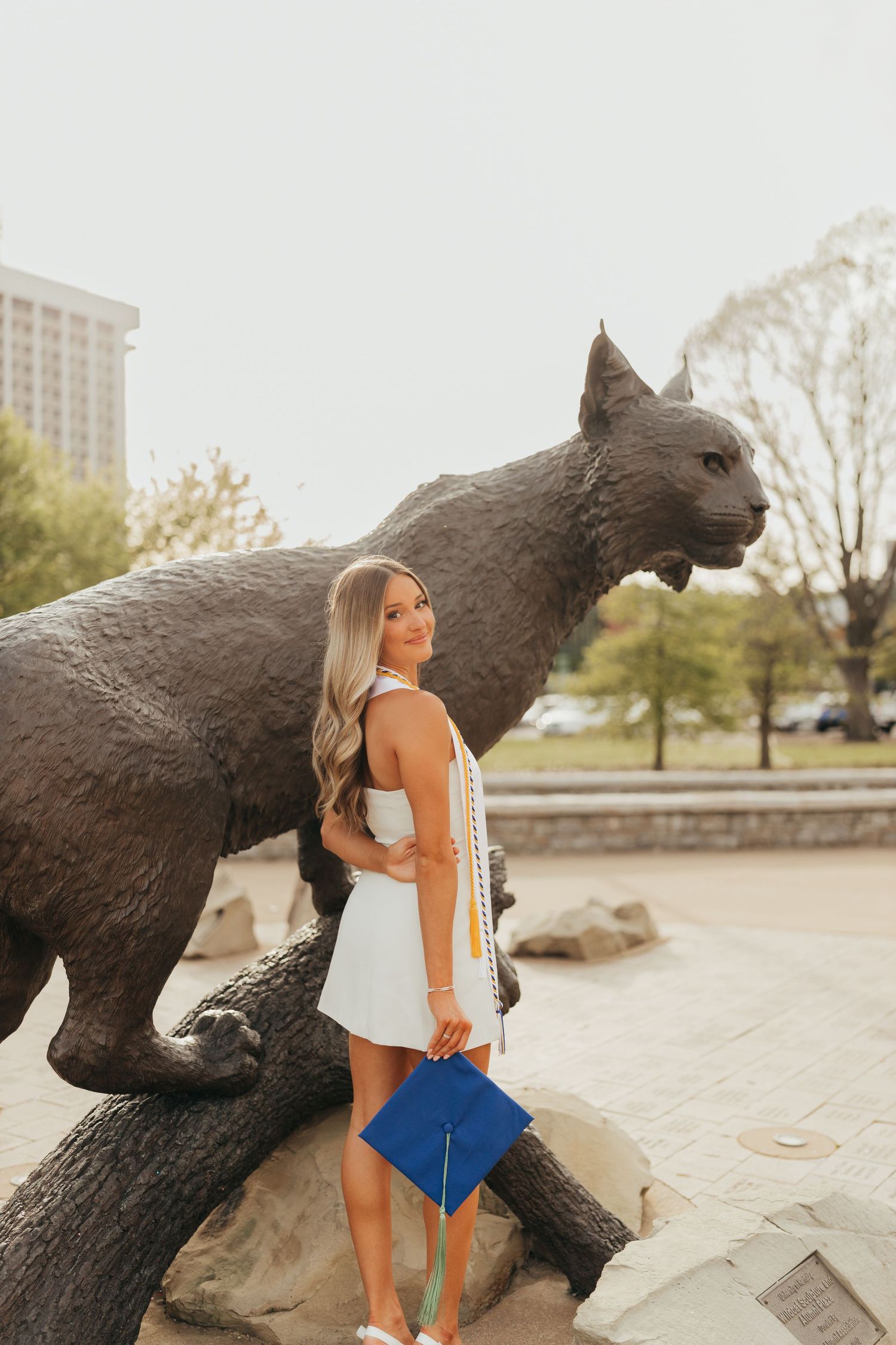 Graduate poses by large wildcat statue while holding blue diploma folder.