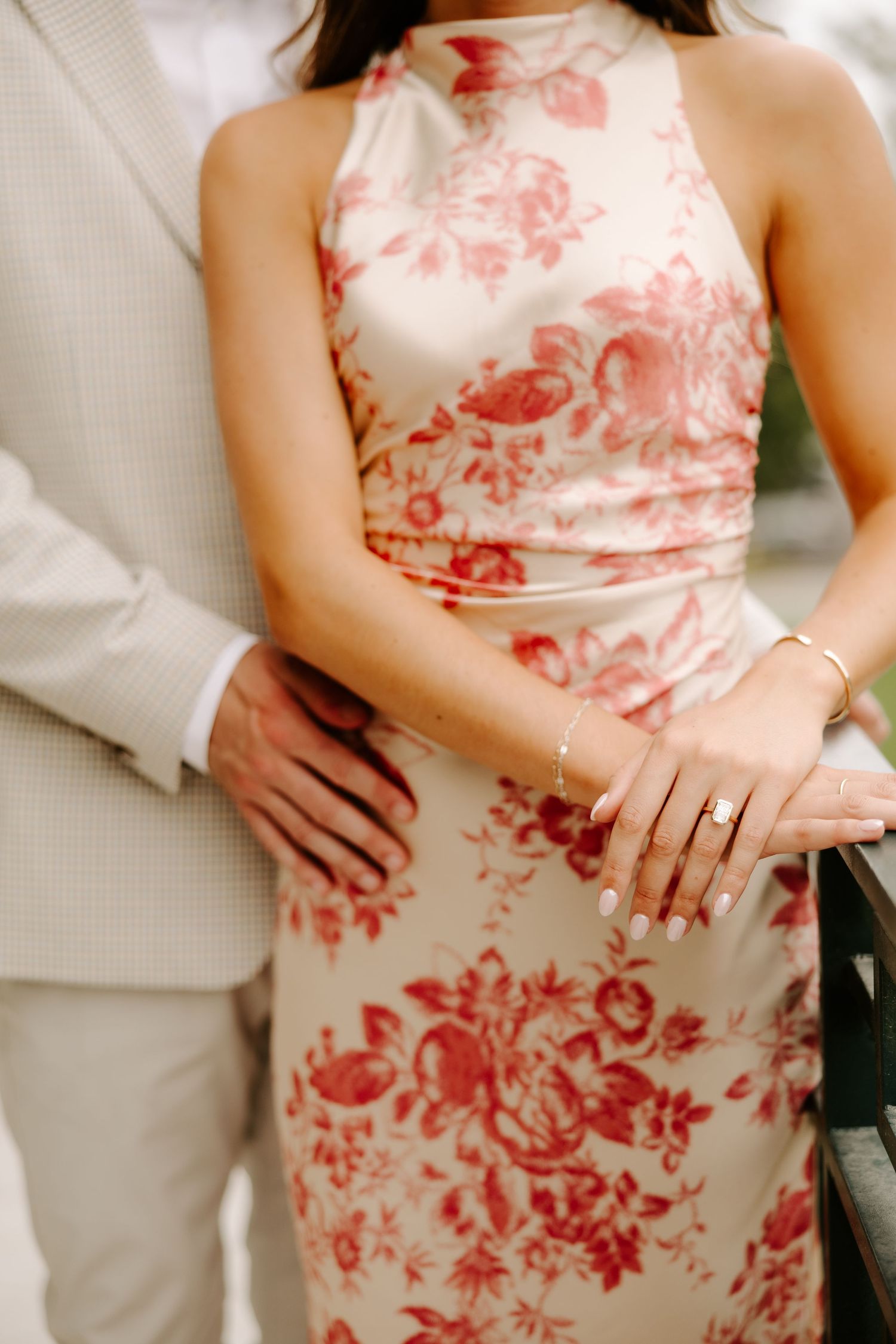 Close-up of a couple embracing, highlighting the womans floral dress, engagement ring, and gold bracelet.