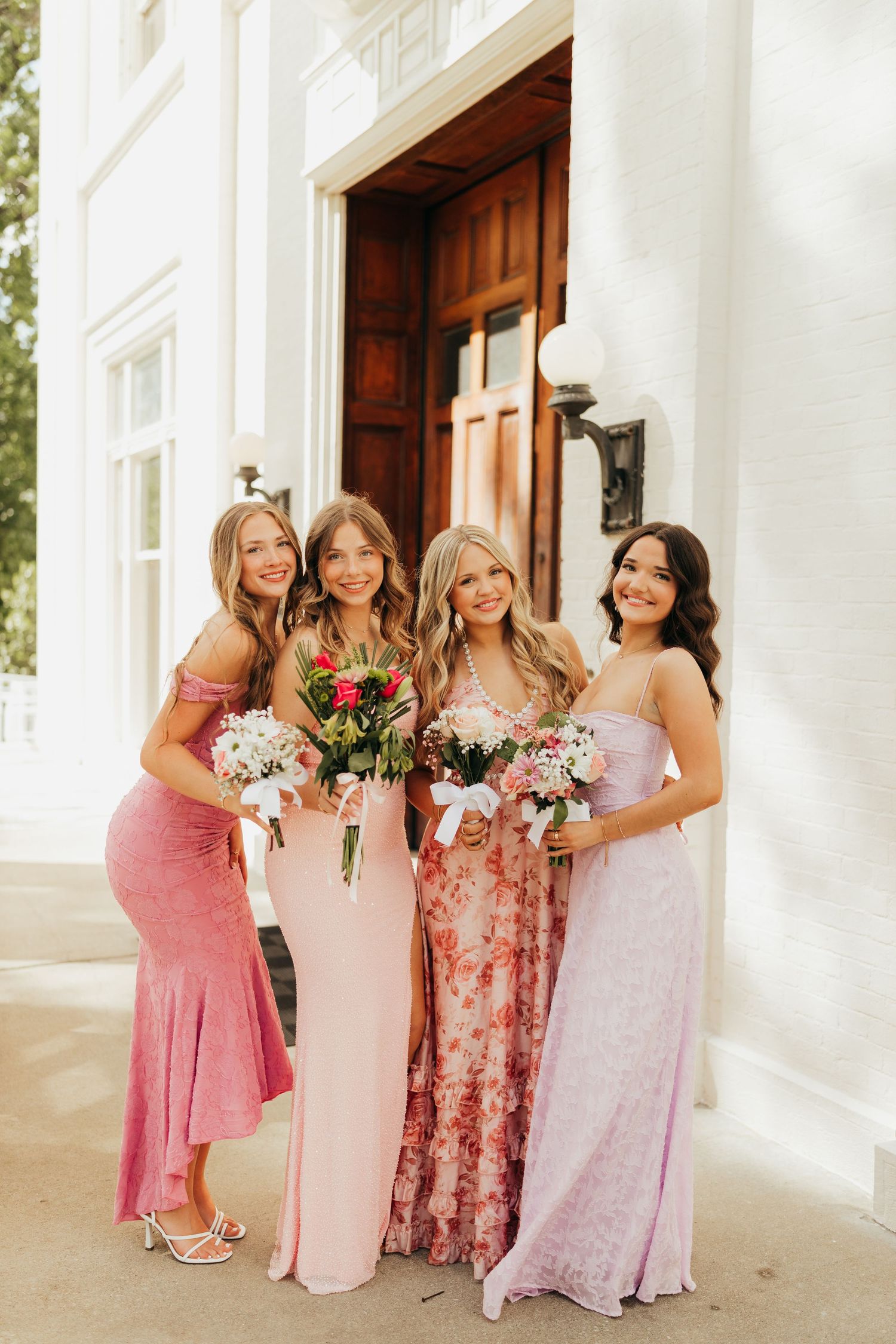 Four young women in pink and floral formal dresses holding bouquets outside a white building entrance.