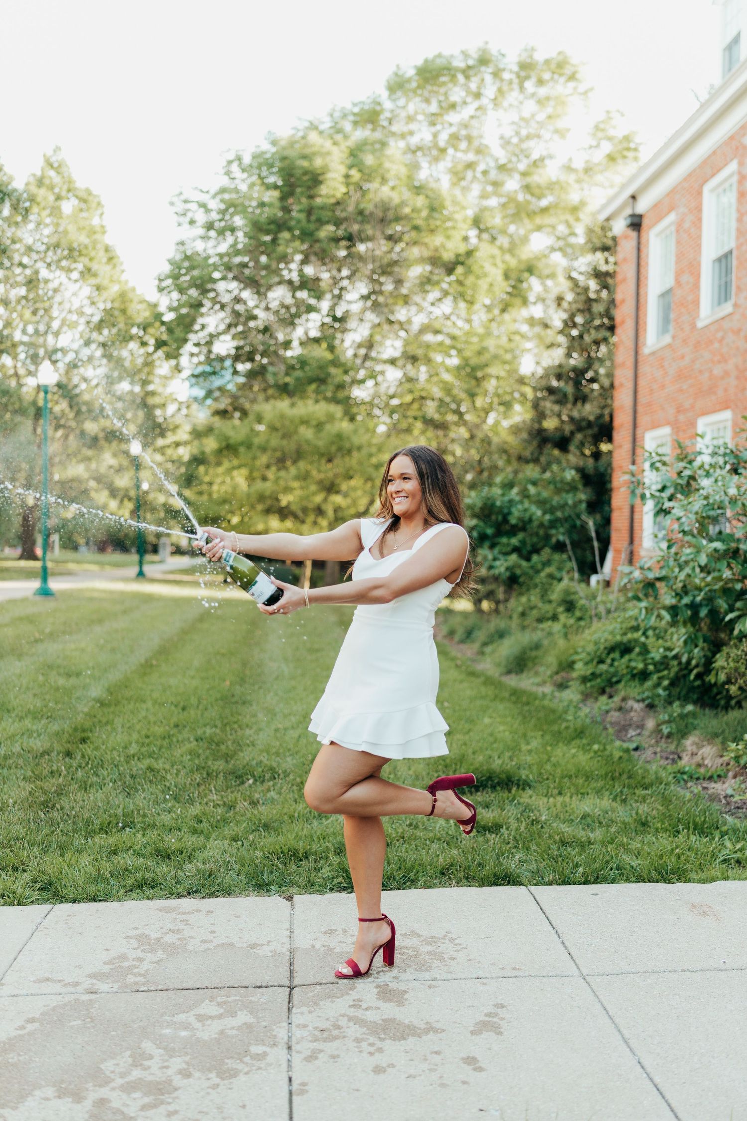 Graduate in white dress celebrates with champagne pop on campus sidewalk.
