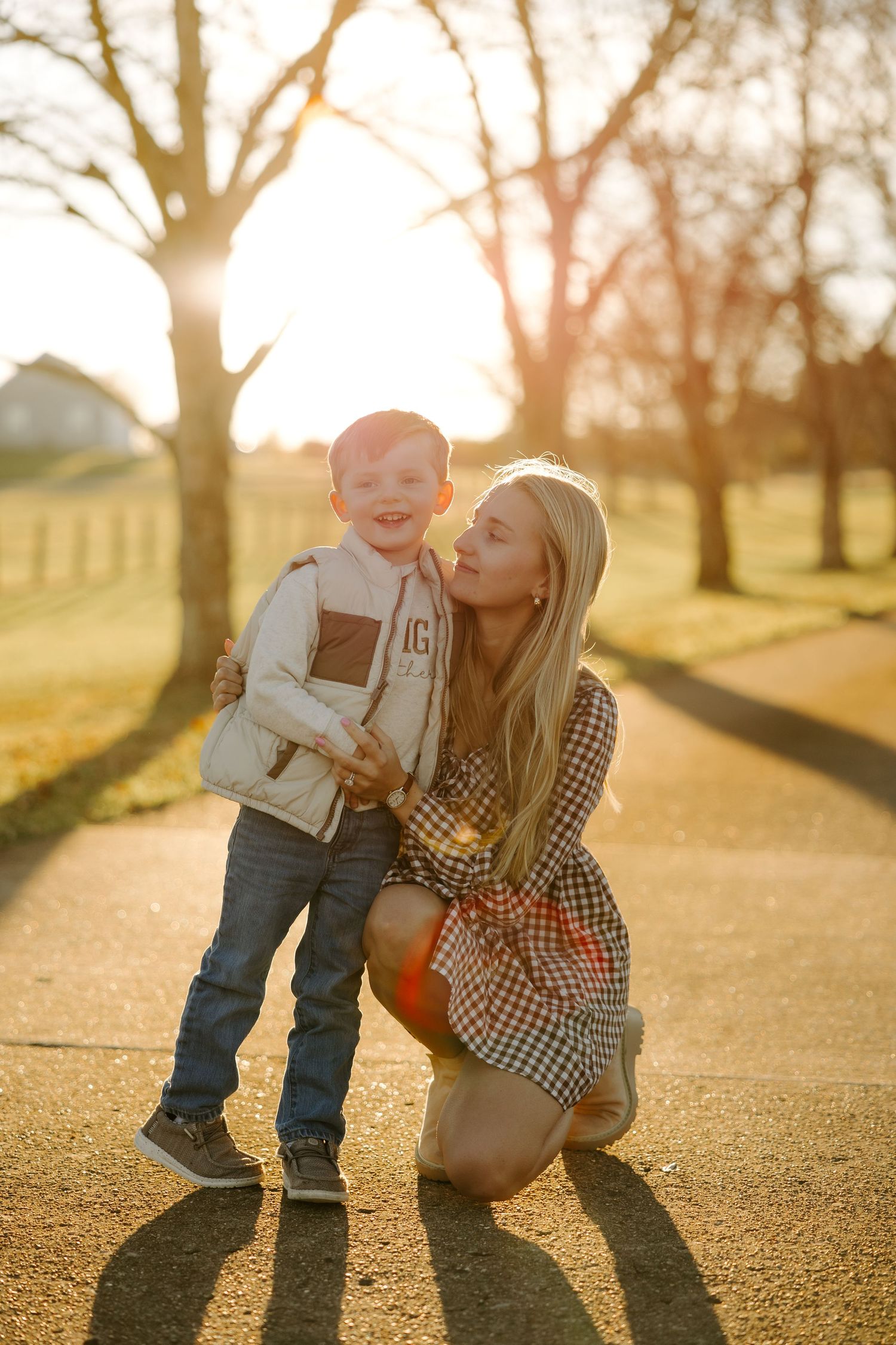 Blonde mother kneeling beside laughing toddler son on a sunlit tree-lined path at golden hour.