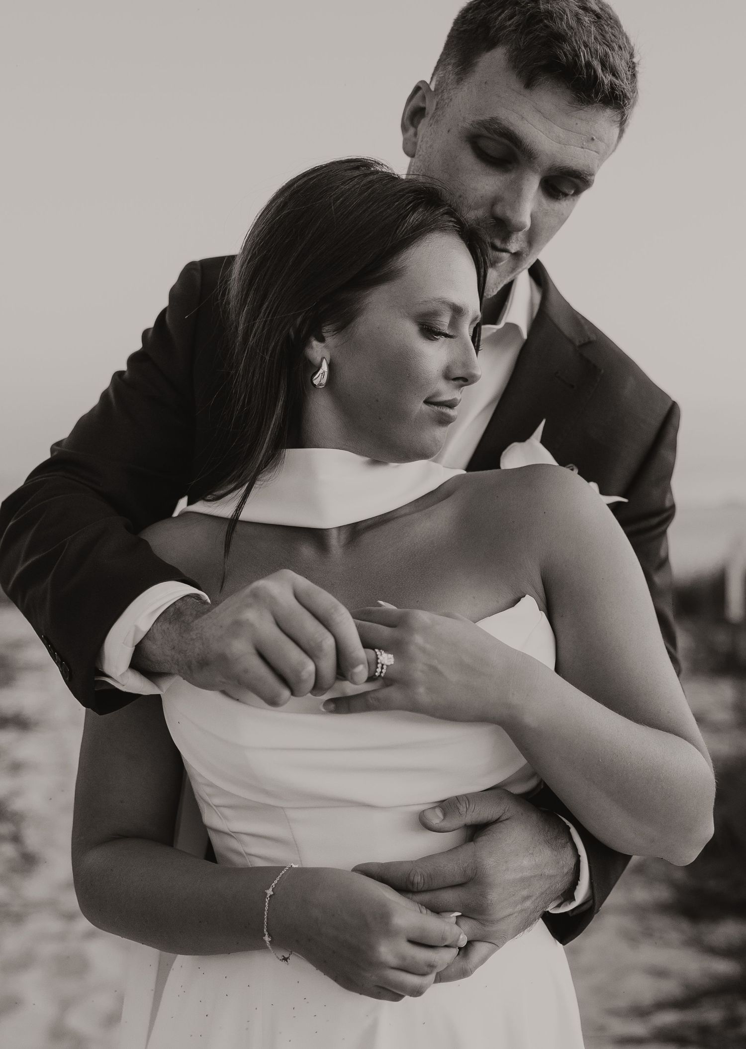 Black and white romantic wedding photo of groom embracing bride from behind in elegant attire.