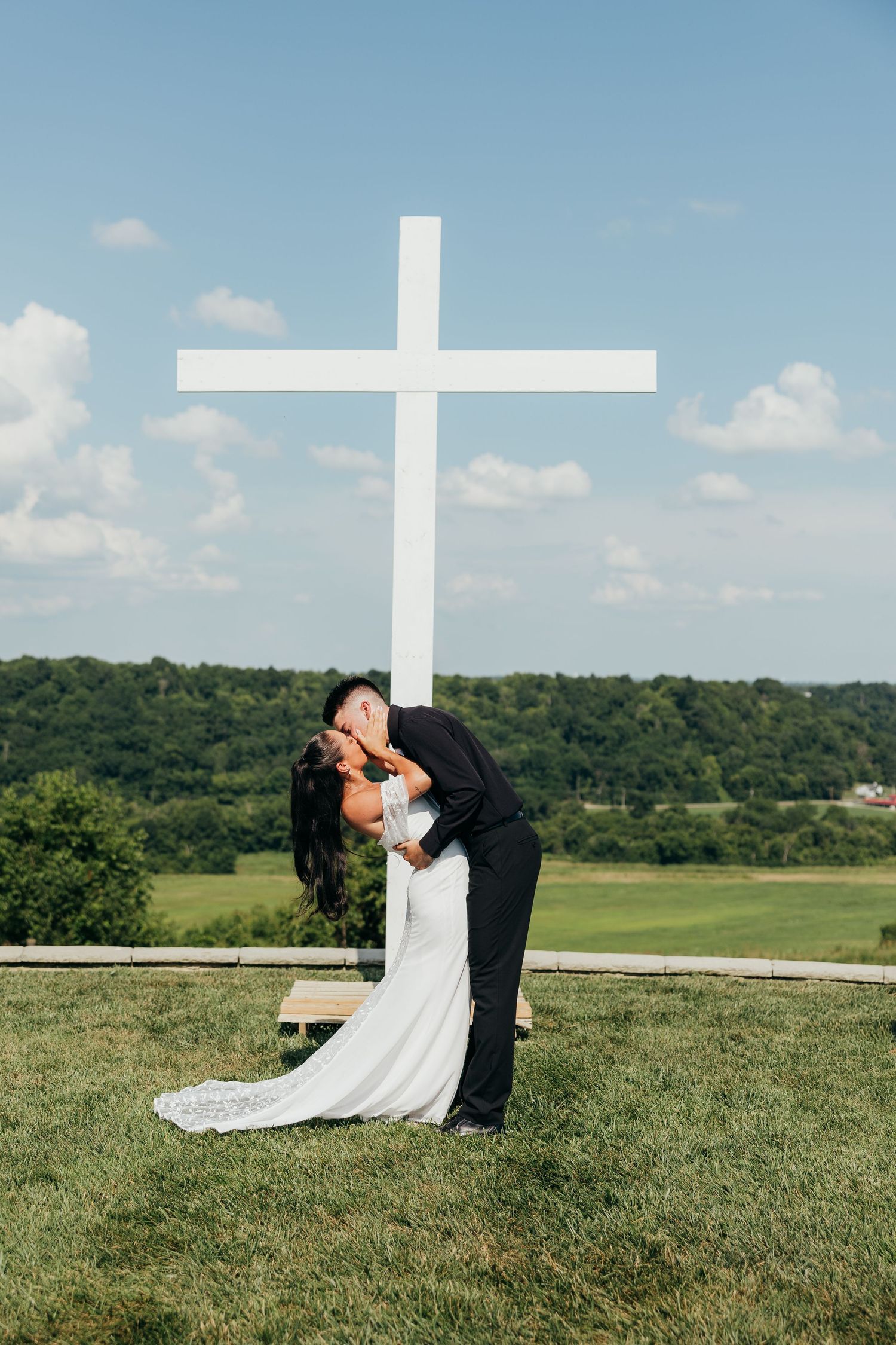 Bride and groom share a romantic dip kiss in front of a large white cross on a green hillside.