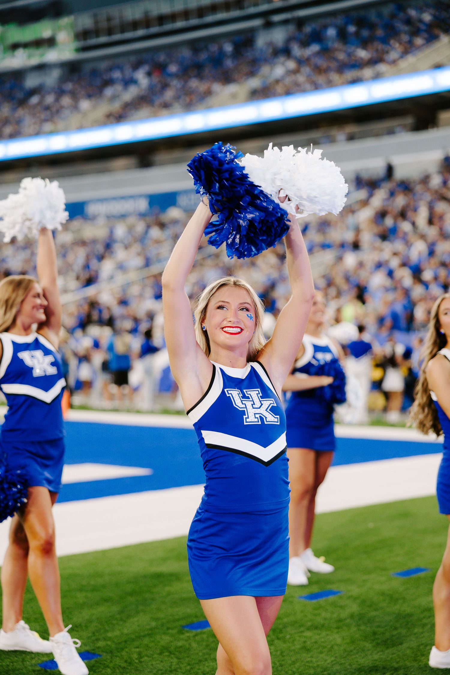 A UK cheerleader in a blue and white uniform raises pom-poms high during a Kentucky Wildcats football game.