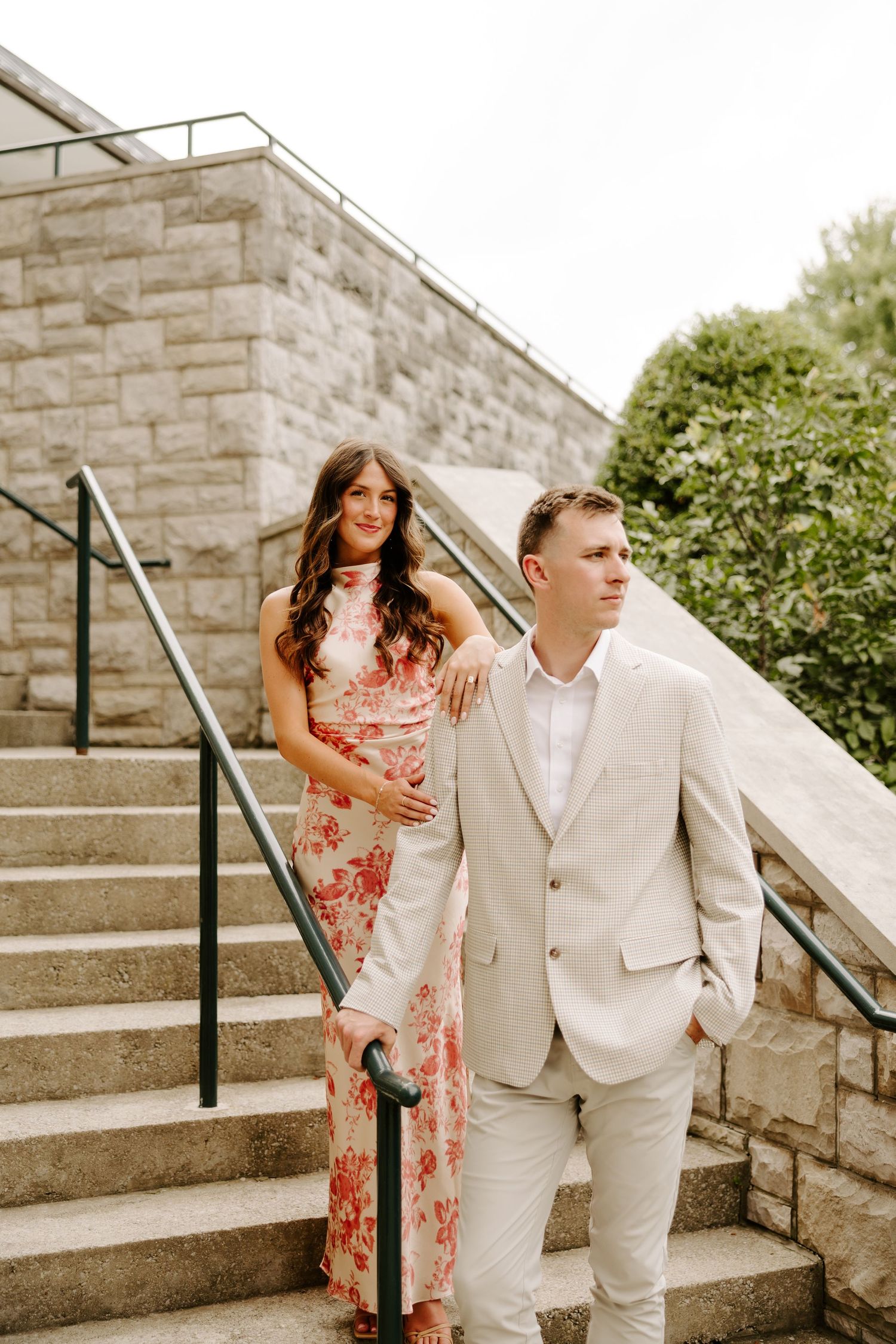 Couple descending stone stairs outdoors, woman in pink floral dress resting hand on mans shoulder.