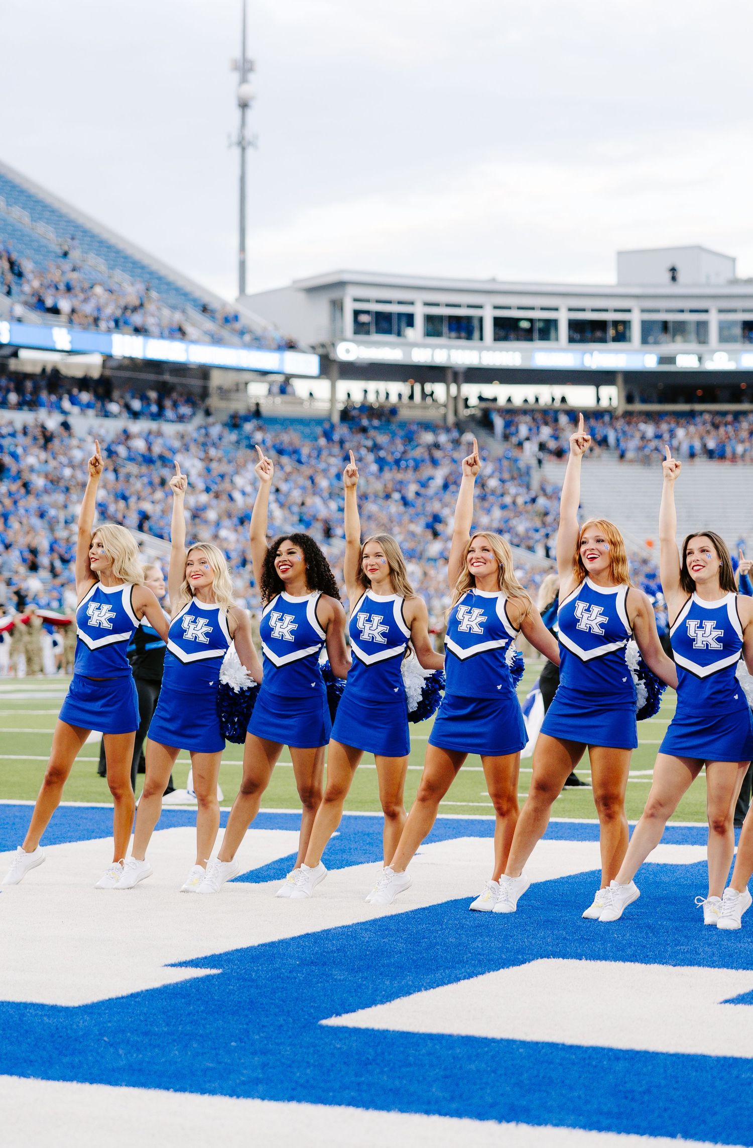 University of Kentucky cheerleaders in blue uniforms performing on field at packed Kroger Field stadium.