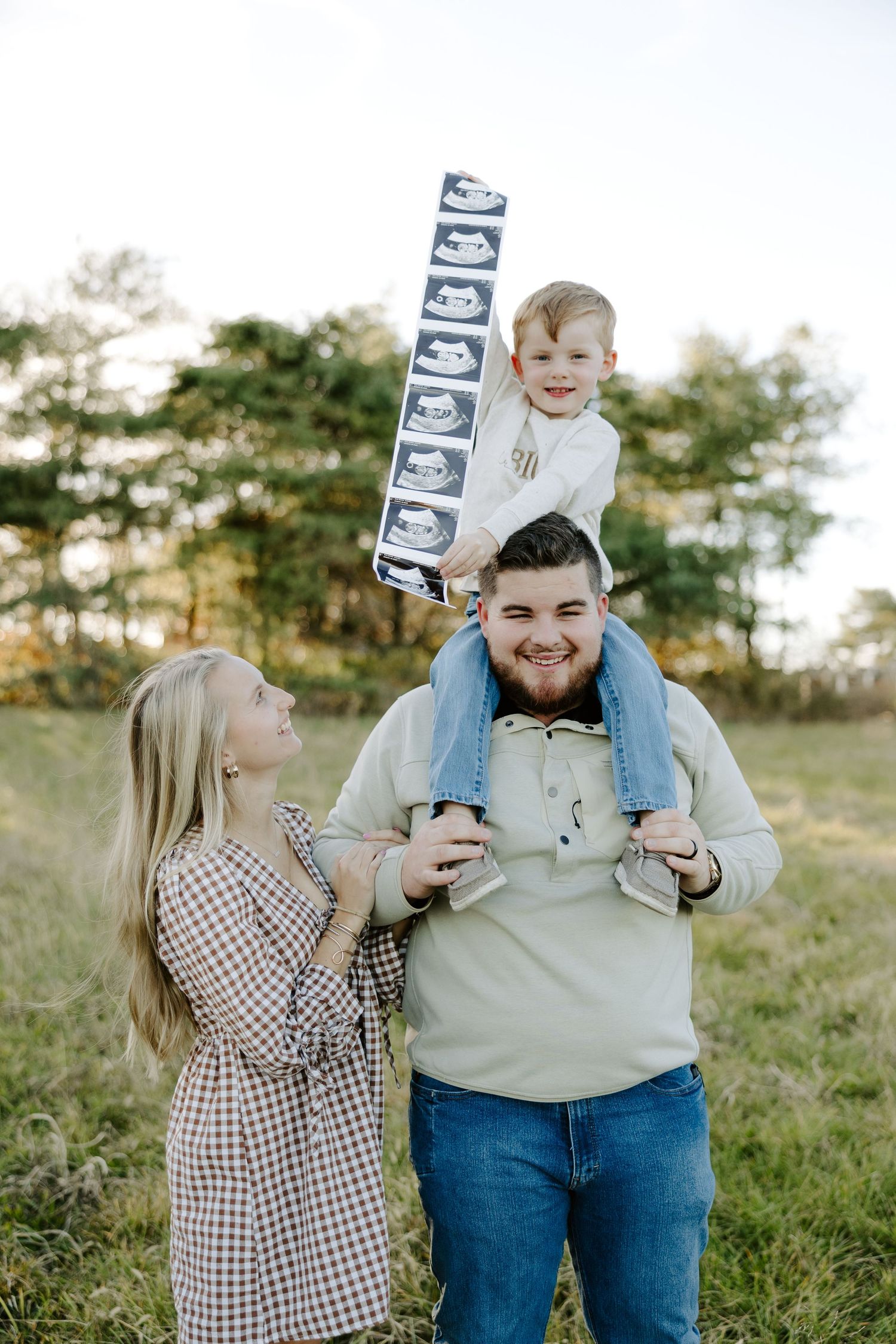 Joyful family photo with young boy on father's shoulders holding ultrasound strip, announcing pregnancy outdoors.