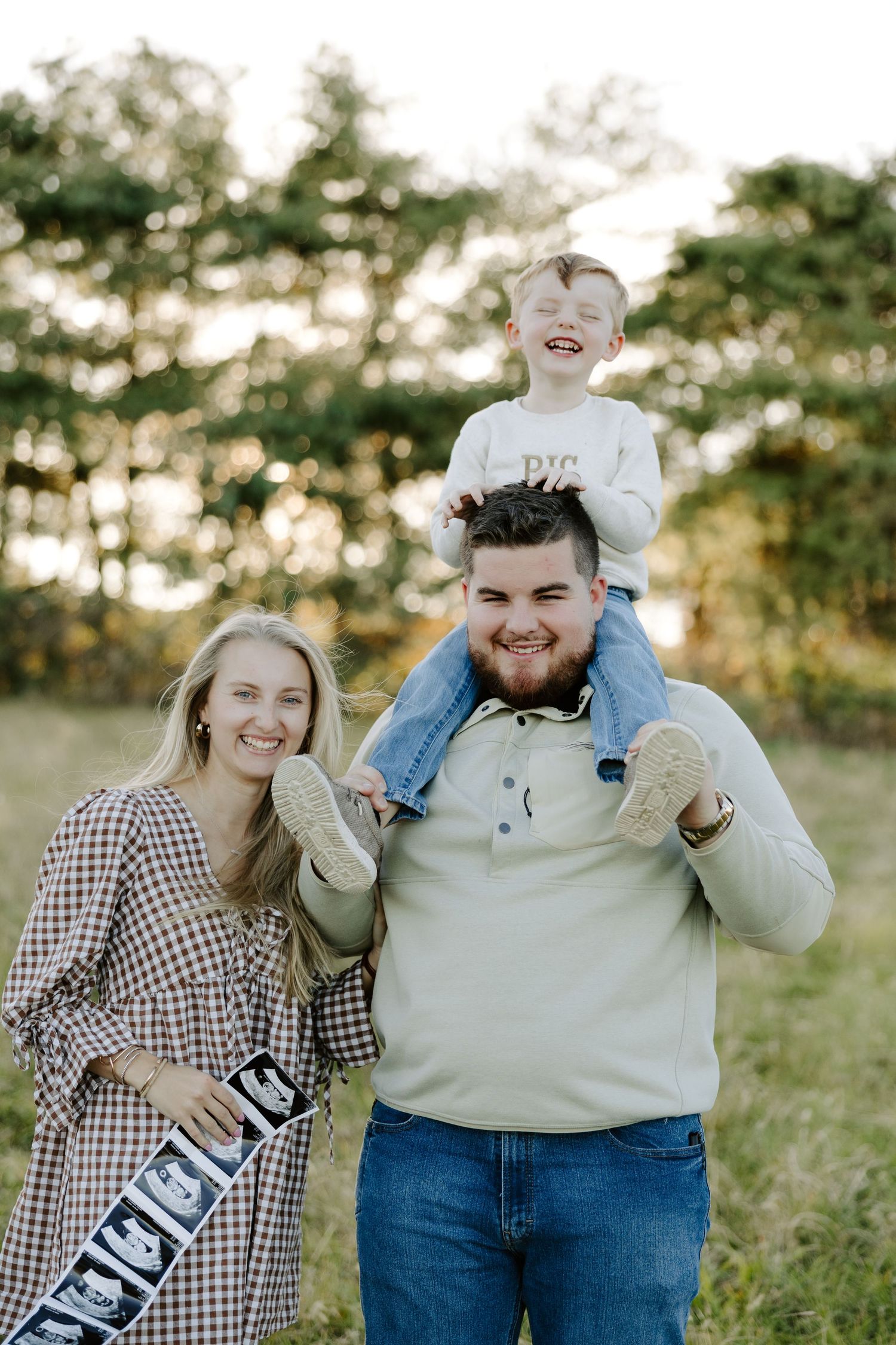 Happy family pregnancy announcement outdoors, parents smiling with toddler on dad's shoulders holding ultrasound photos.