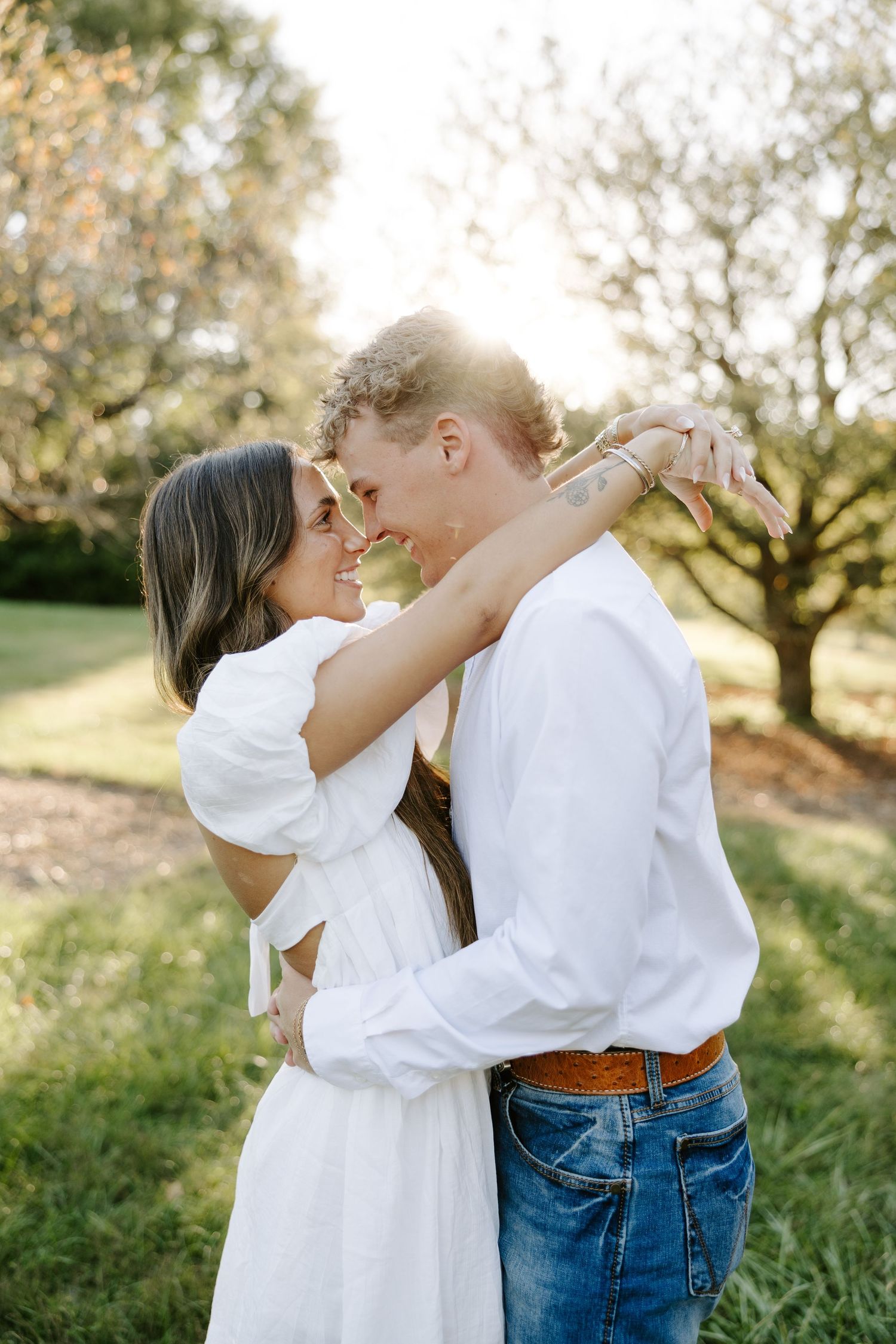 Romantic couple nearly kissing in golden sunlight, woman in white dress with arms around man's neck.