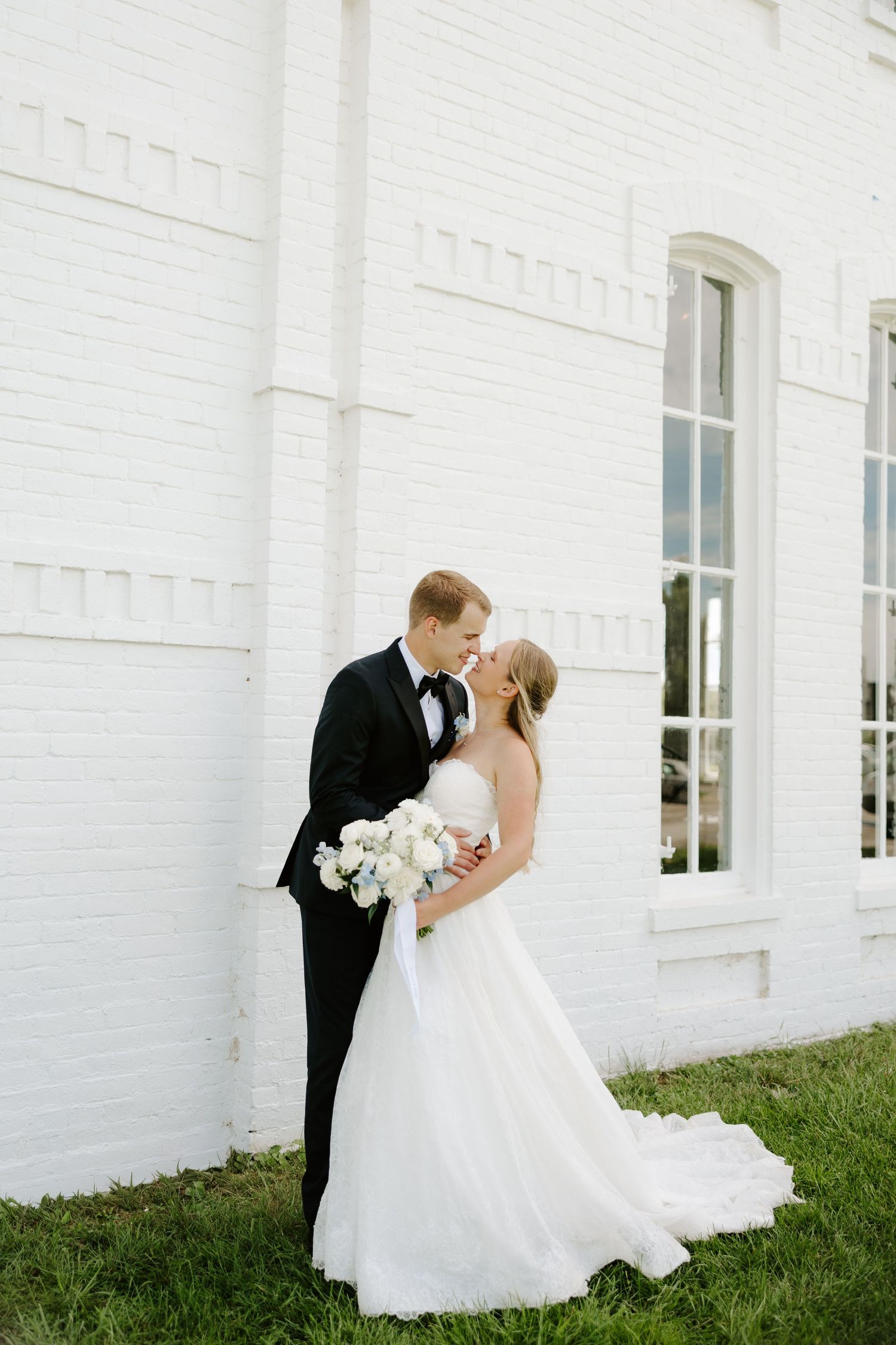 A bride and groom share a kiss beside a white brick building, bride holding a white bouquet.