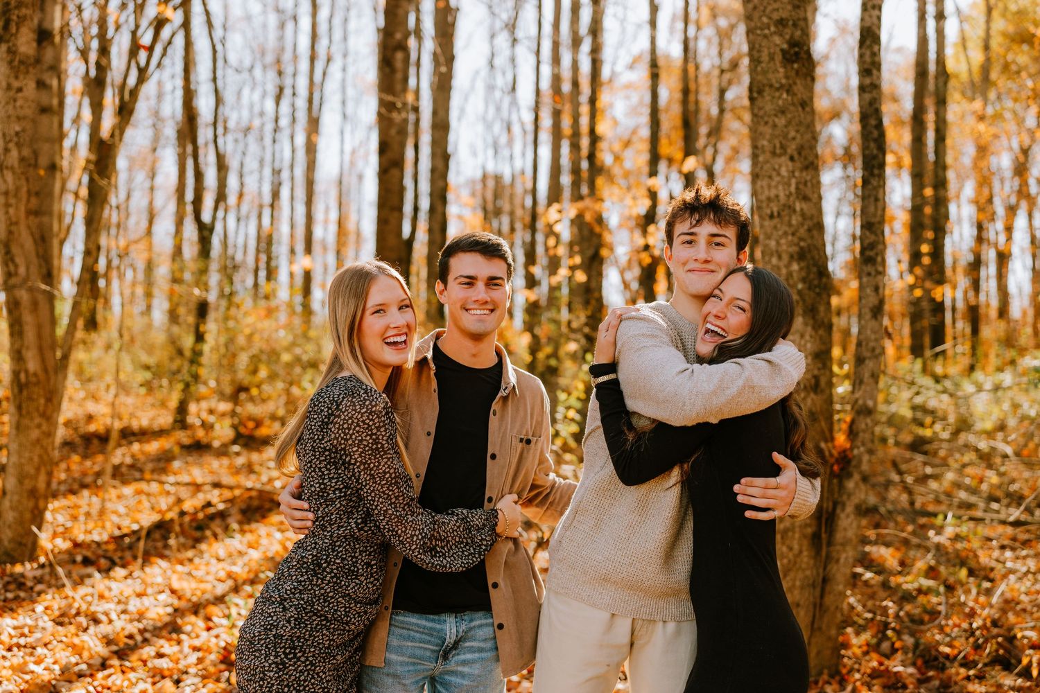 Two couples laughing and hugging each other in a sunlit autumn forest with golden leaves.