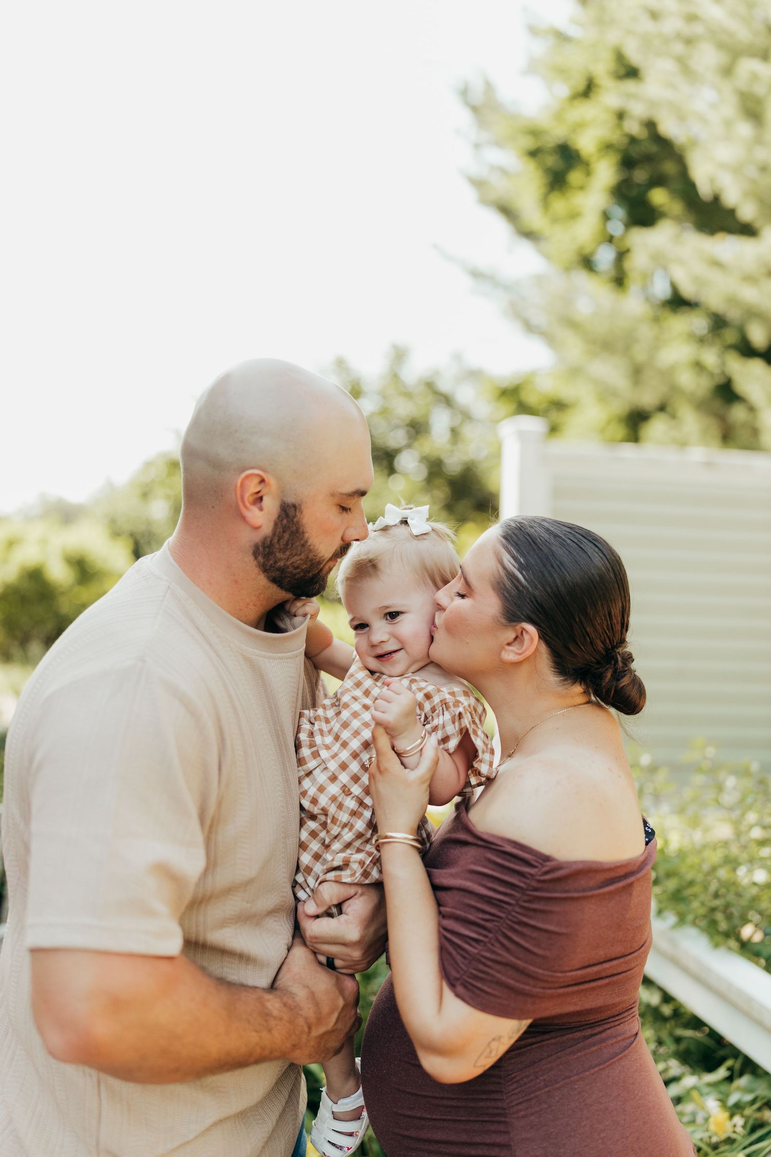 Parents kissing their smiling toddler daughter on cheeks during outdoor maternity family portrait.