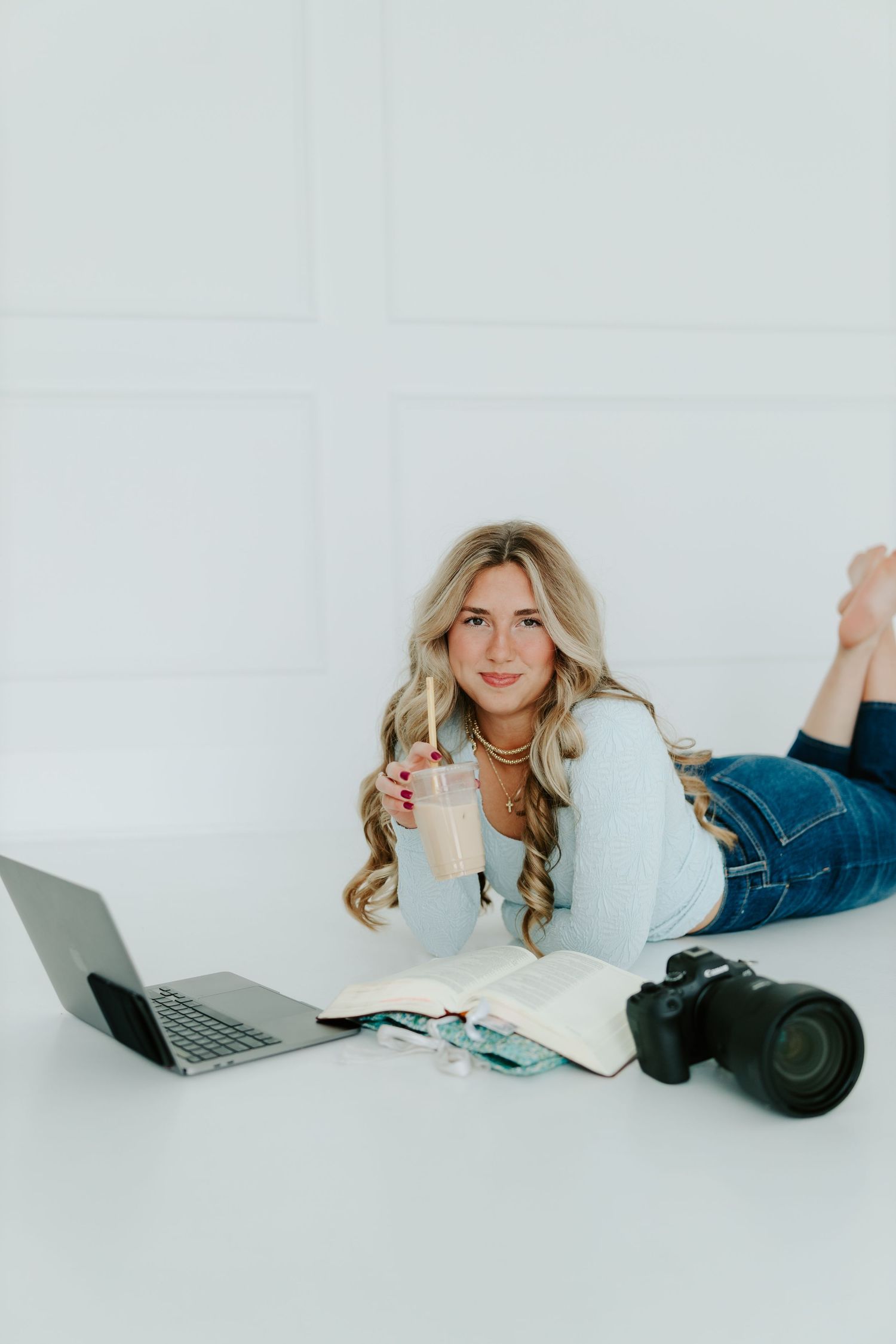 Blonde woman lying on floor smiling, holding iced drink beside laptop, open book, and DSLR camera.