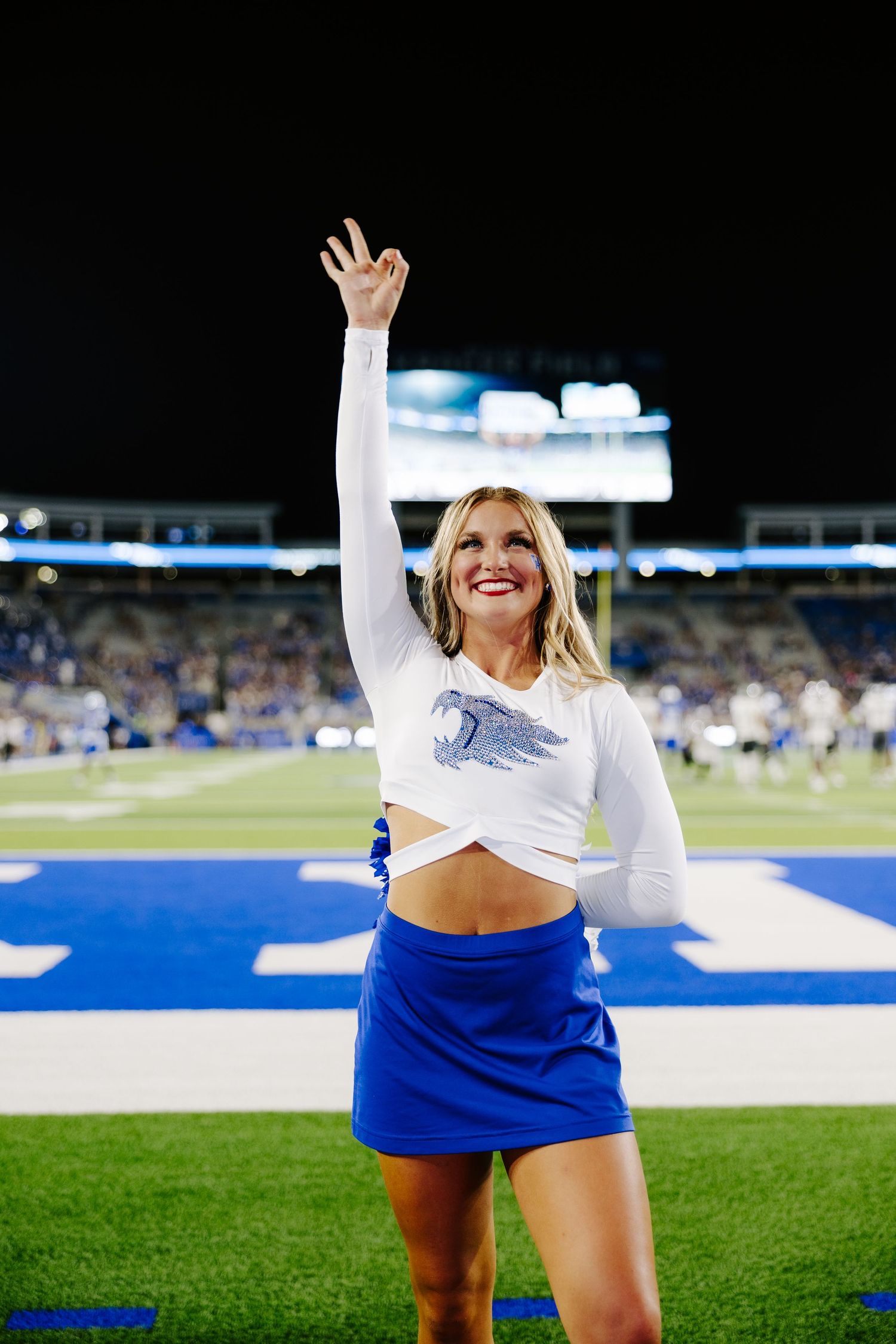 Smiling blonde Kentucky Wildcats cheerleader in white crop top and blue skirt waving at night game.