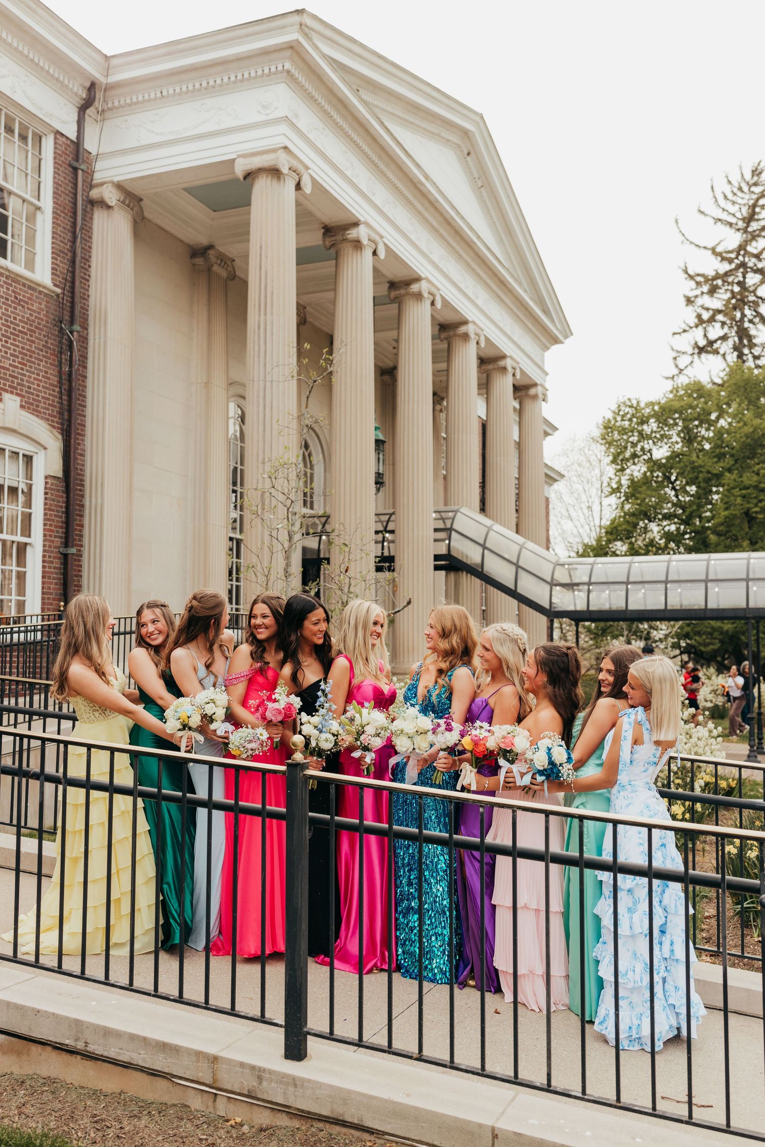 Group of women in rainbow-colored formal dresses holding bouquets outside a neoclassical brick building.