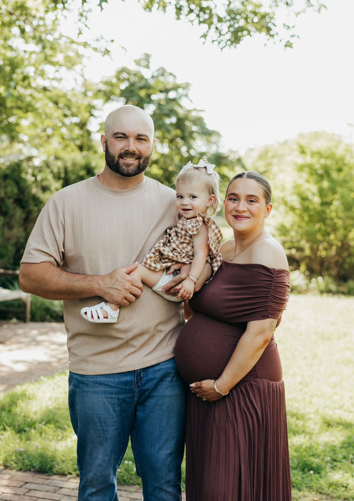 Happy family maternity photo with bald dad holding baby girl and pregnant mom in burgundy dress outdoors.