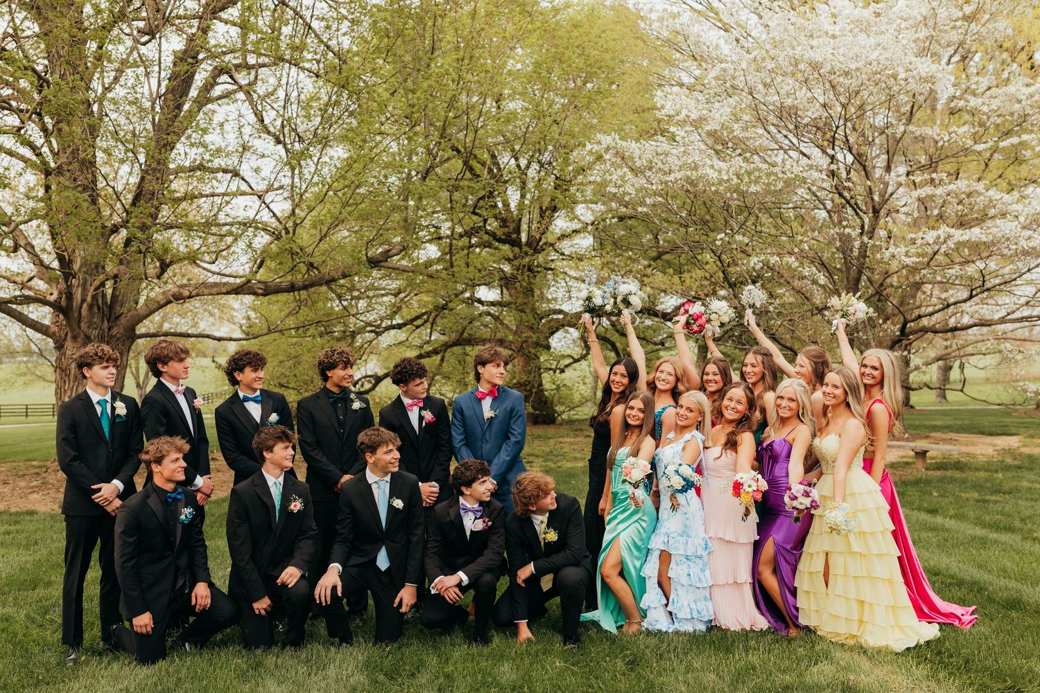 Large prom group of teens in formal wear posing outdoors on green grass with blooming trees.