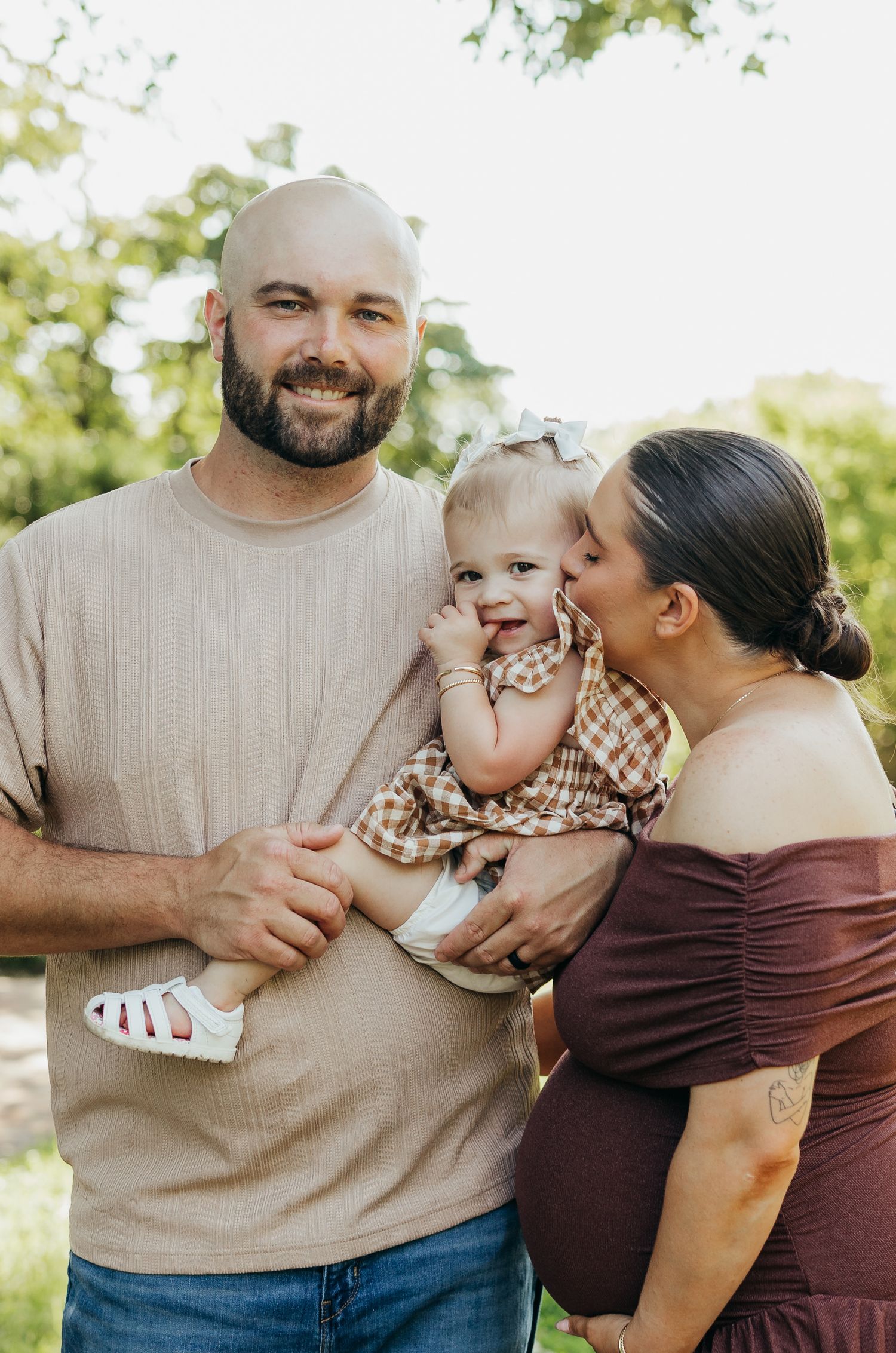 Loving family portrait with dad holding baby while pregnant mom kisses childs cheek in green park.