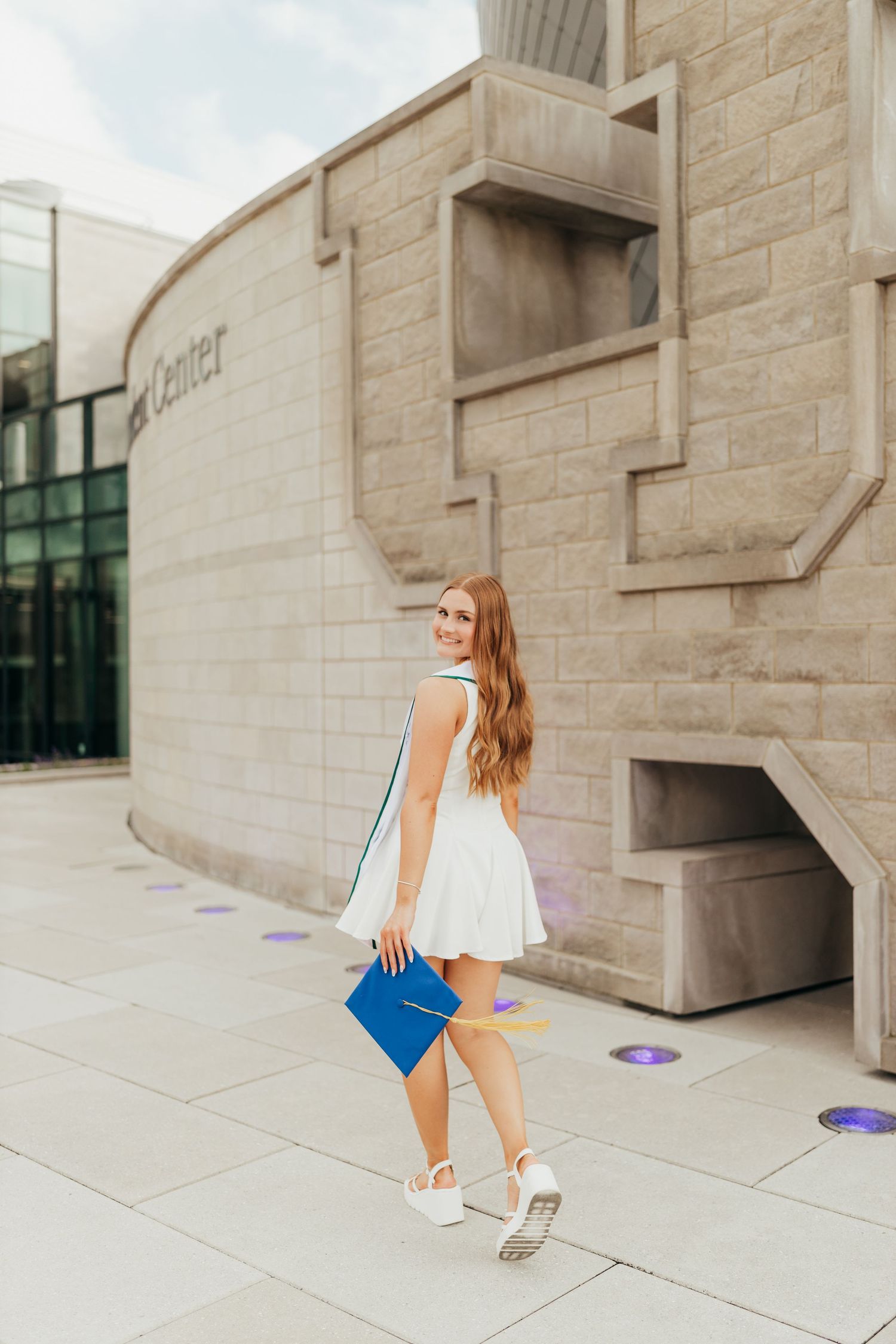 Young woman in white dress holding blue graduation cap walking outside a stone university building.