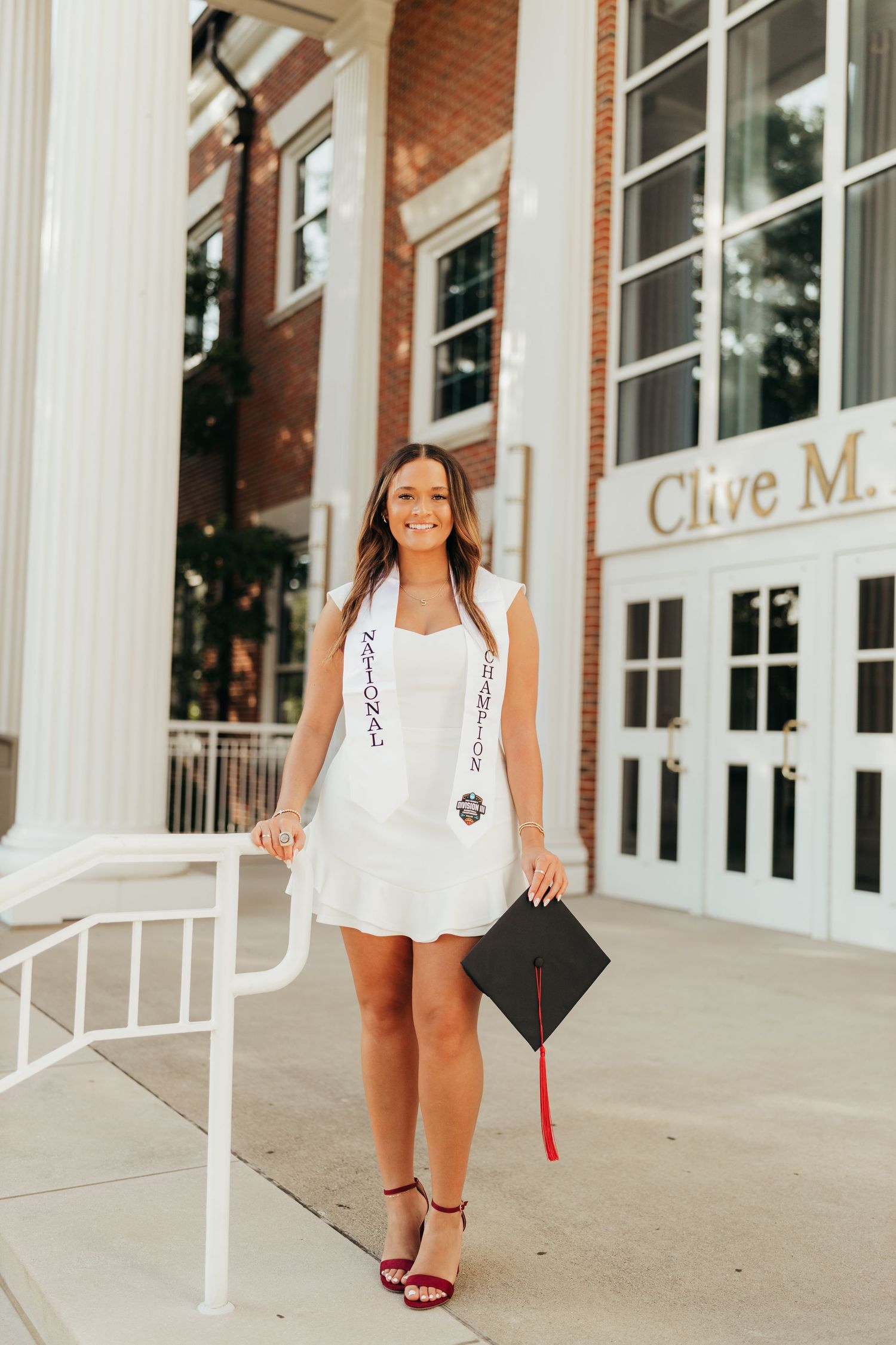 Graduate in white dress holds graduation cap near brick building with columns.