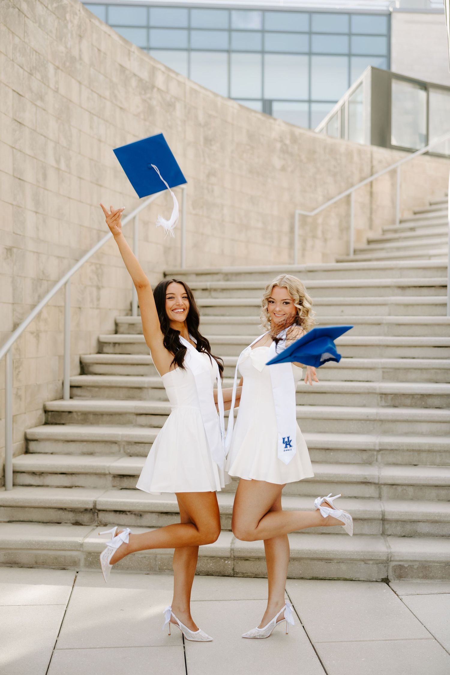 Two women in white dresses celebrate graduation by tossing blue caps on outdoor stone steps.