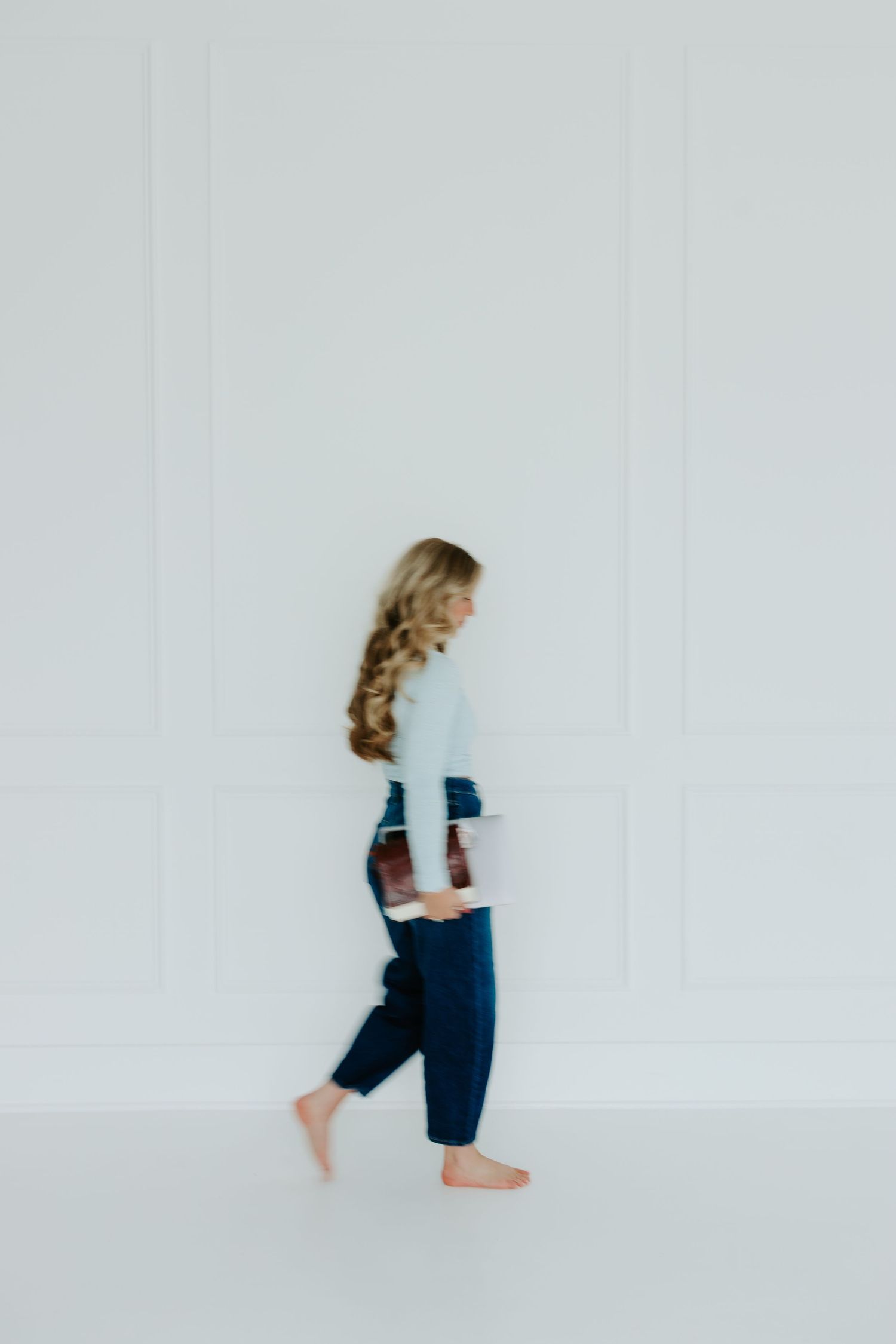 Blurred motion photo of blonde woman walking barefoot in blue jeans and light top against white wall.