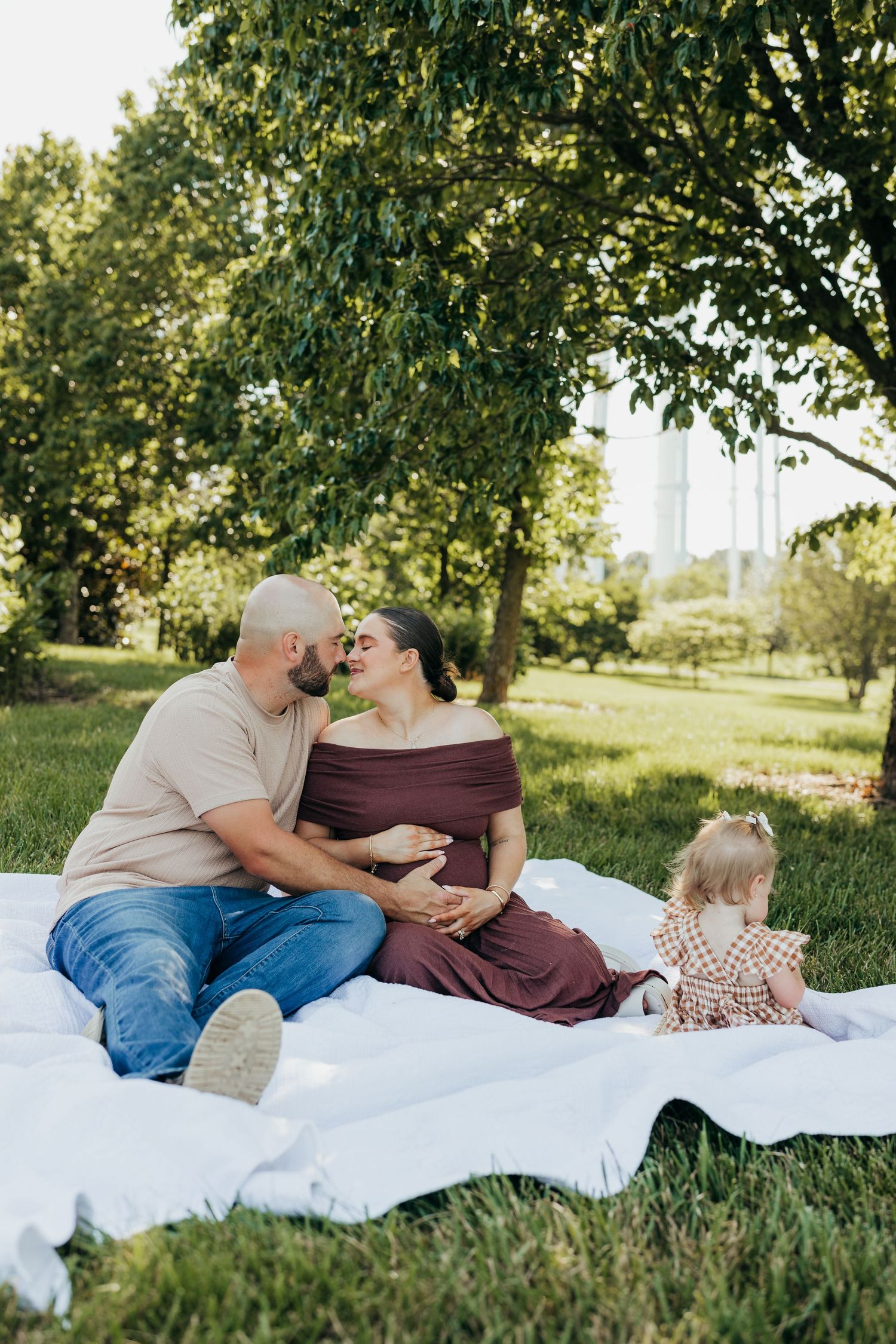 Expecting couple sharing a kiss on white blanket in park while toddler plays beside them.