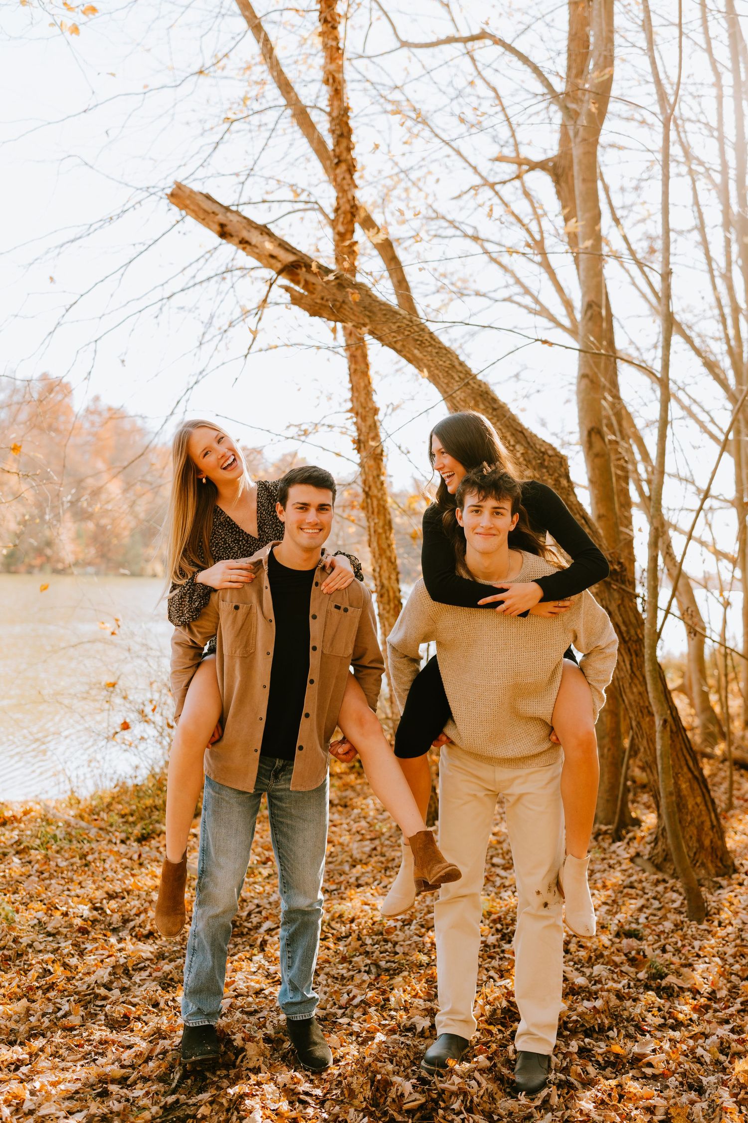 Two young men giving piggyback rides to two young women in a fall forest near a lake.