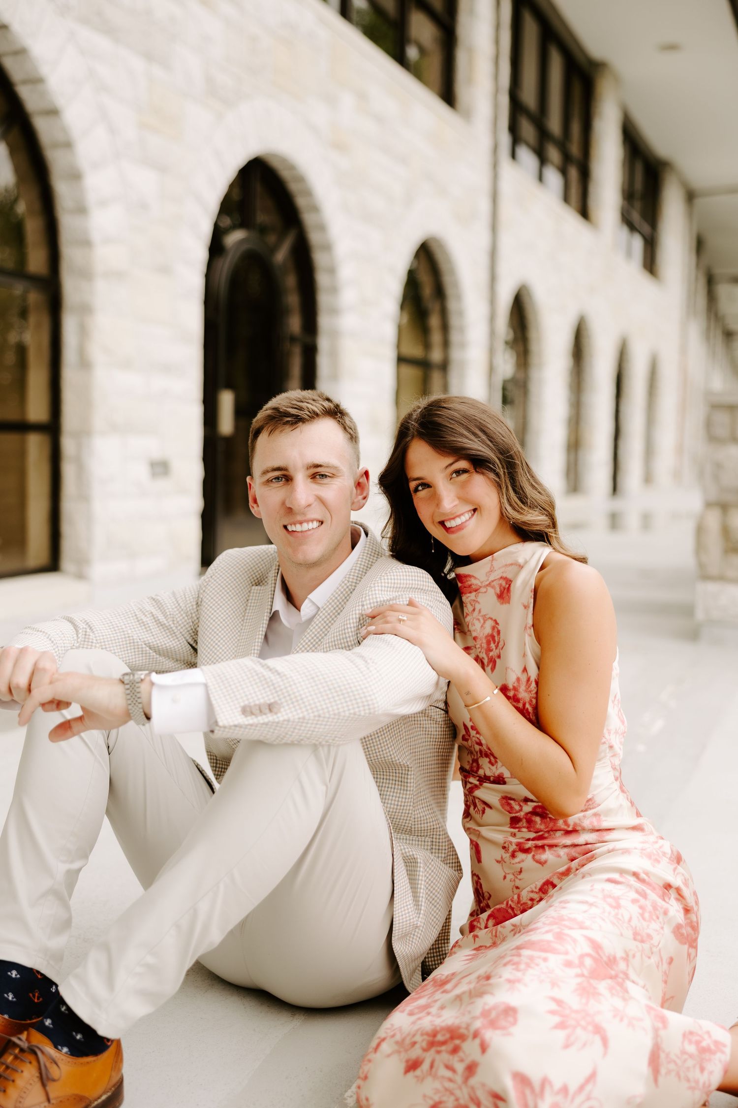 Cheerful couple sitting together in front of a stone building with arched windows, smiling at camera.
