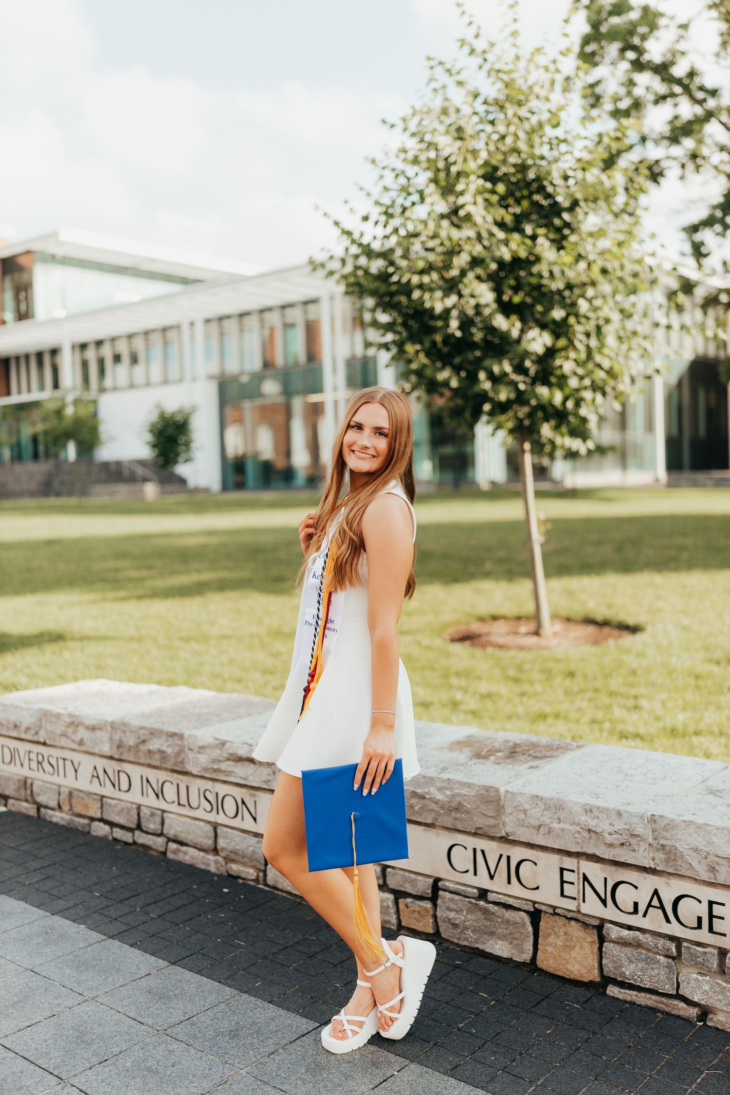 Graduate in white dress with honor stoles holding blue cap near Civic Engagement university sign.