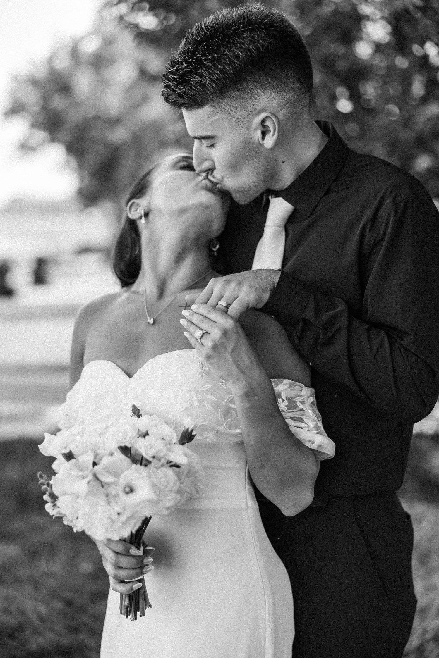 Black and white photo of groom kissing bride on cheek while she holds white wedding bouquet.