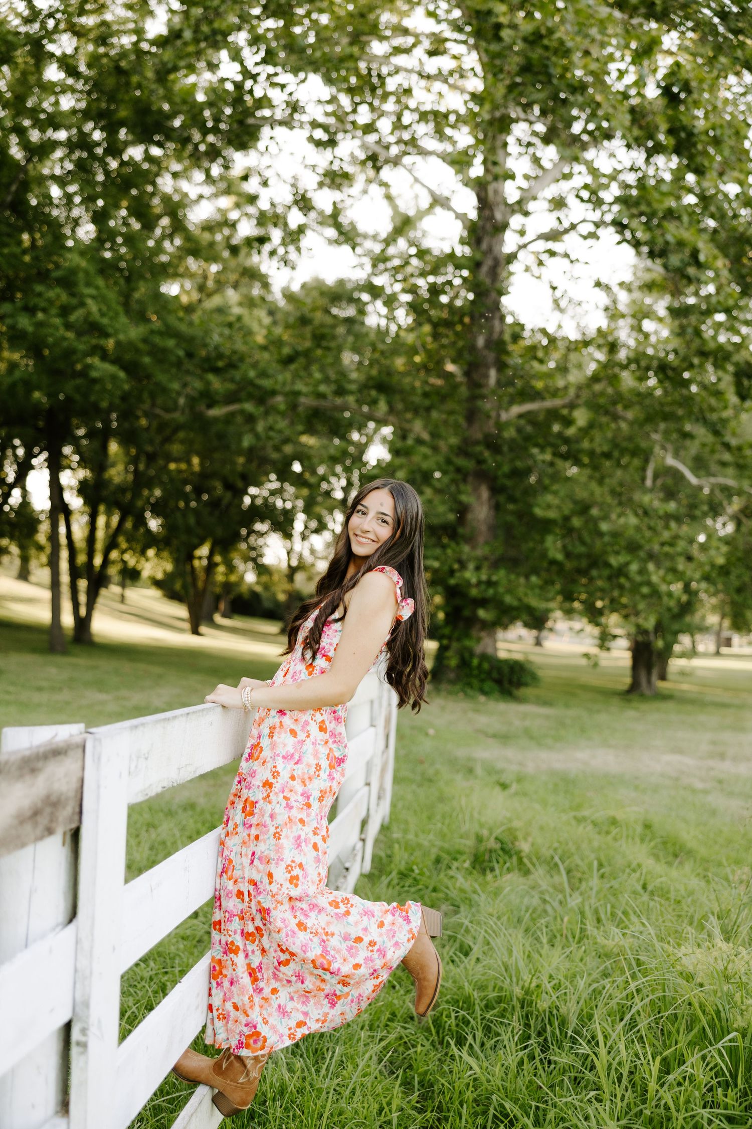 Brunette teen girl in pink floral dress leaning on white fence surrounded by green trees.