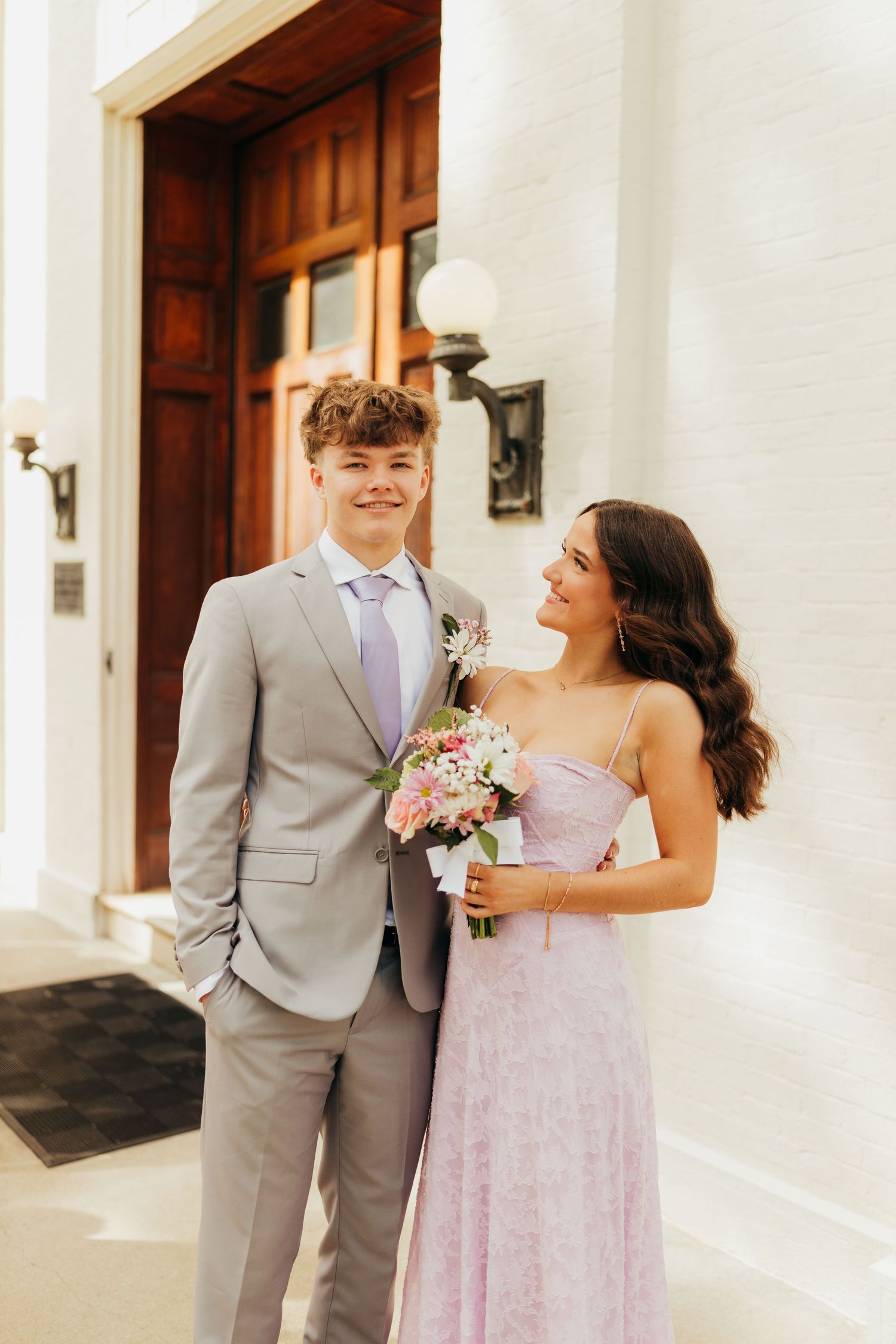 Young couple in prom attire, woman in lavender gown holding bouquet, posing outside a white brick building.