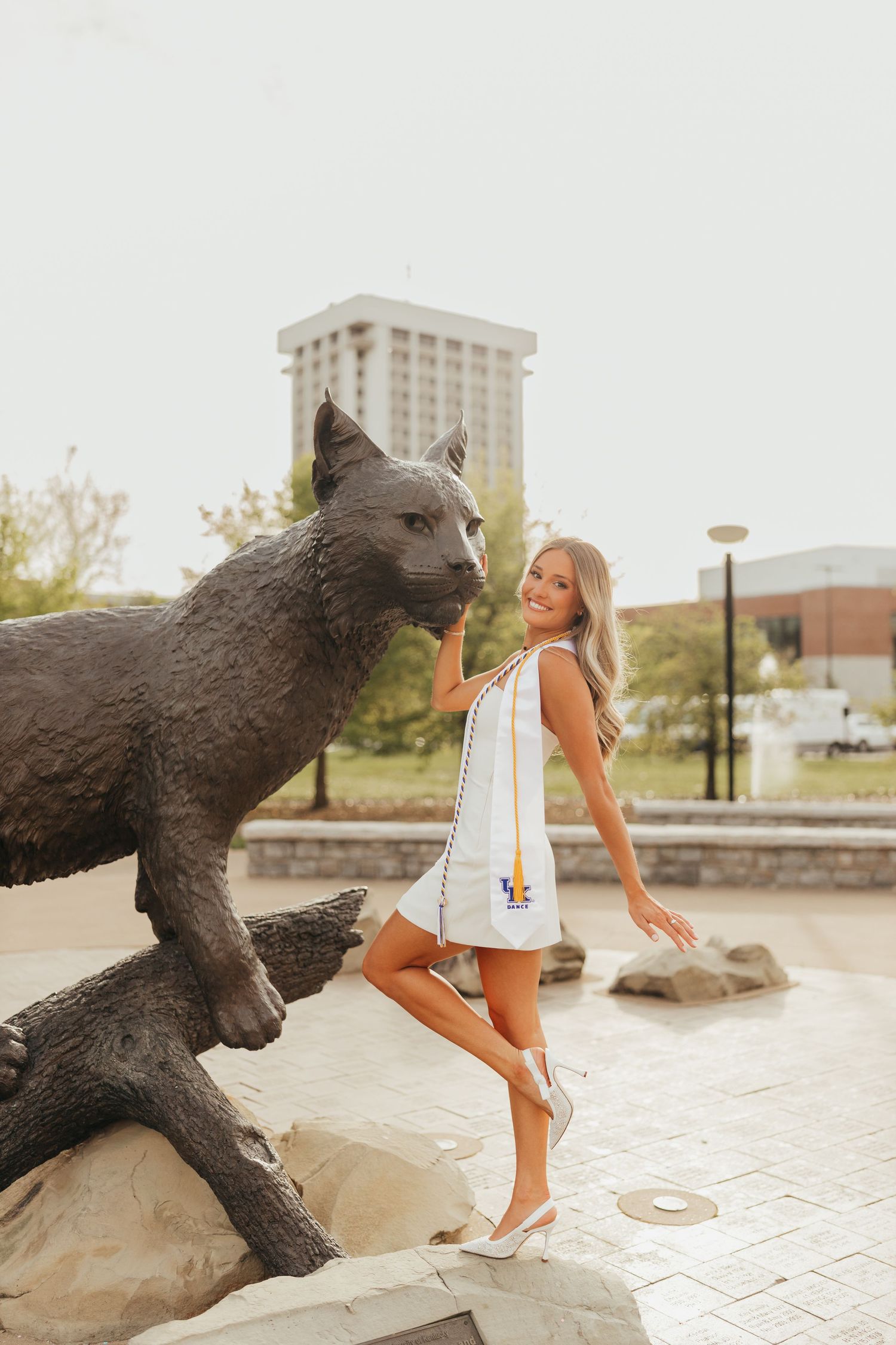 Graduate in white dress strikes playful pose next to large bronze wildcat sculpture.