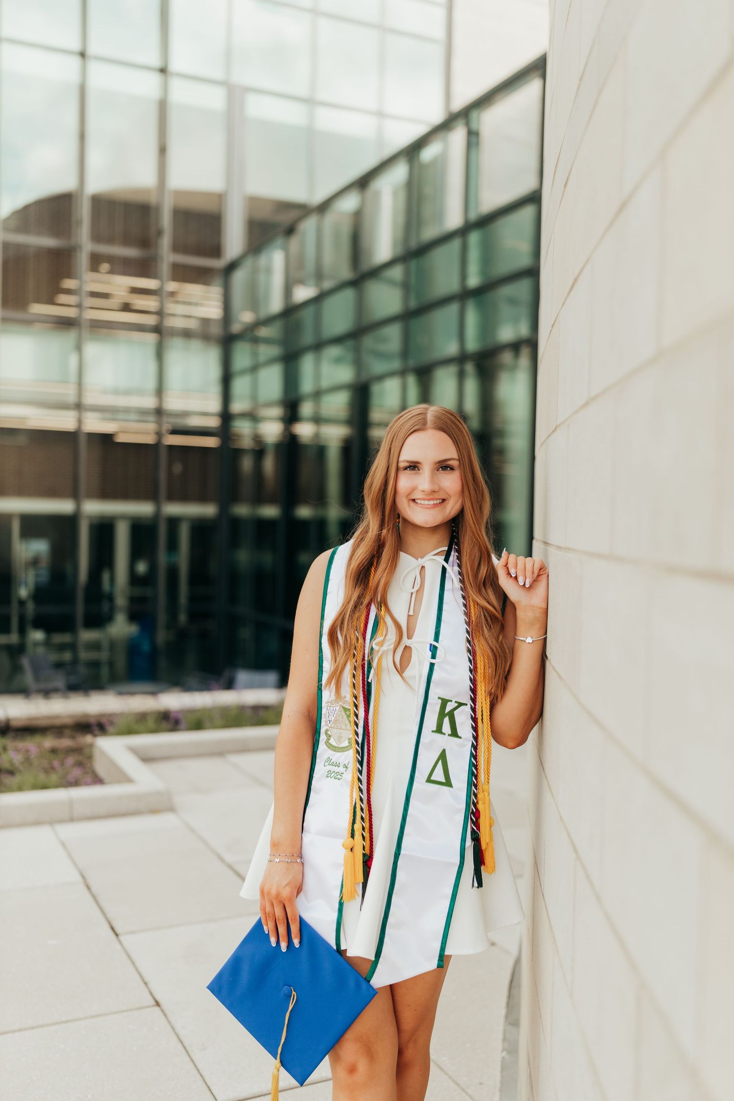 Smiling graduate in white dress with Kappa Delta stole holding blue cap outside glass building.