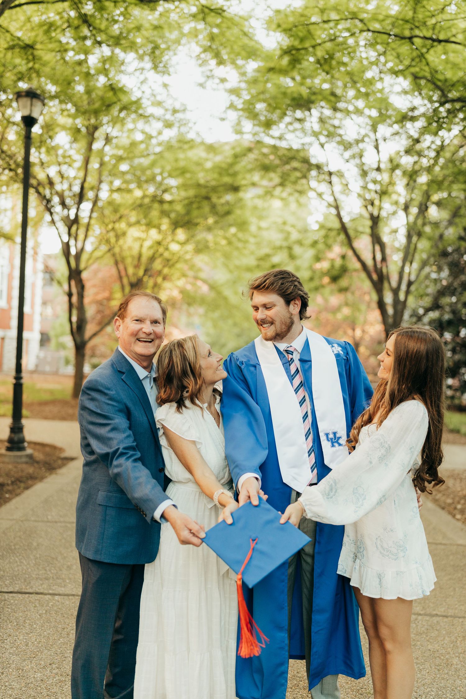 University of Kentucky graduate in blue cap and gown celebrating with family on tree-lined campus path.