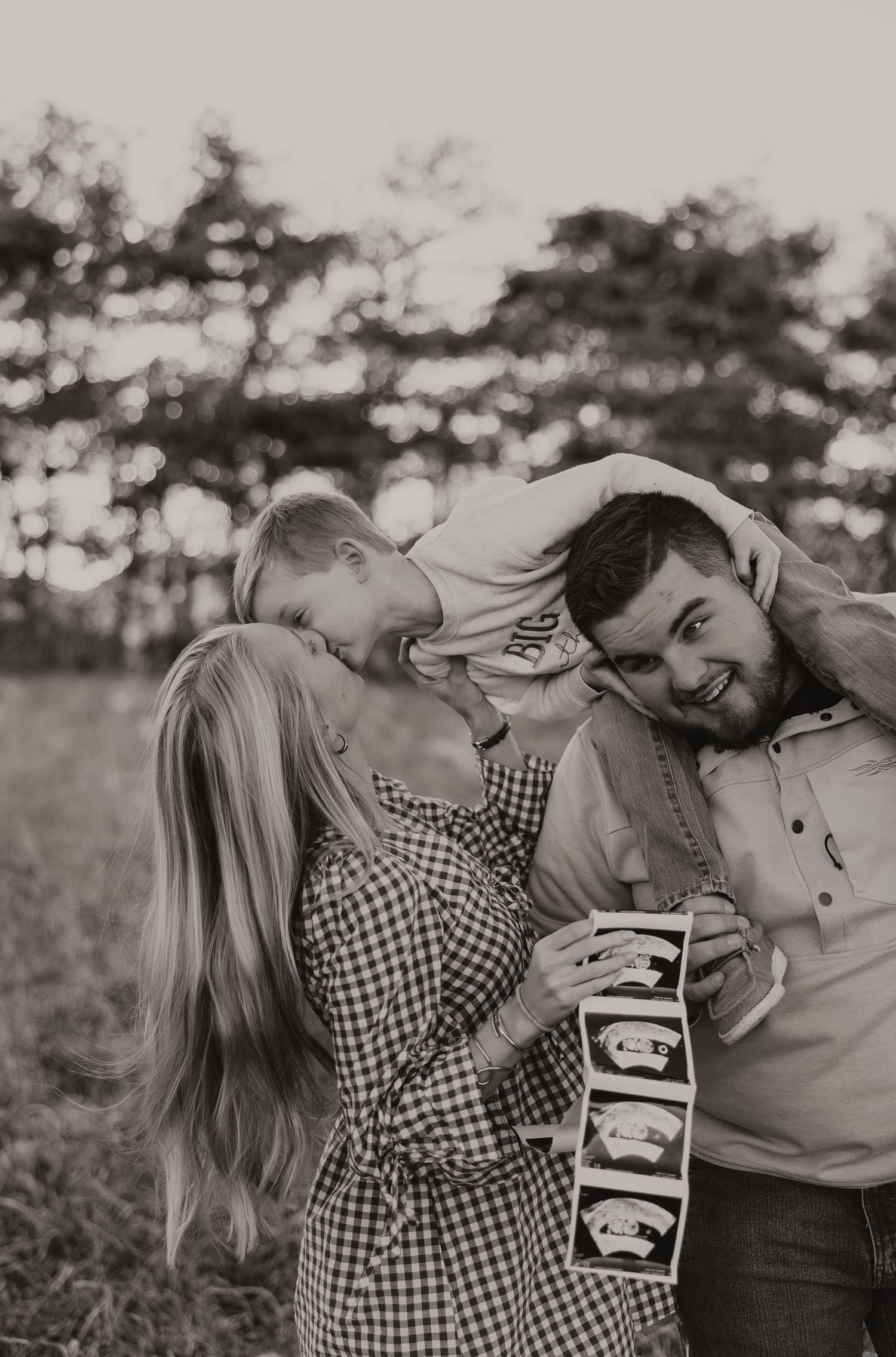 Black and white family pregnancy reveal photo with toddler hugging parents while mom holds ultrasound images.
