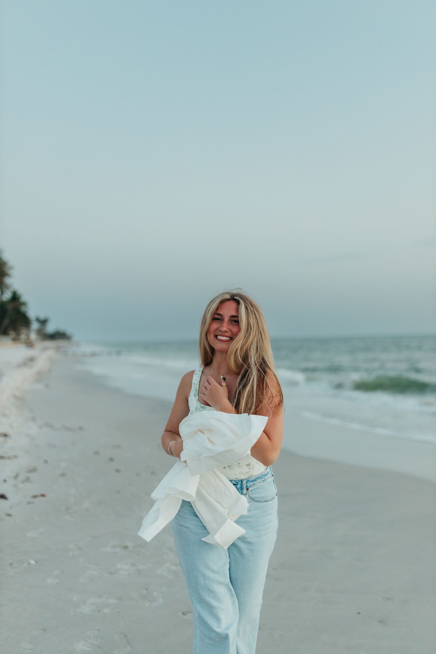 Happy blonde woman in white top and jeans walking along a serene sandy beach at dusk.