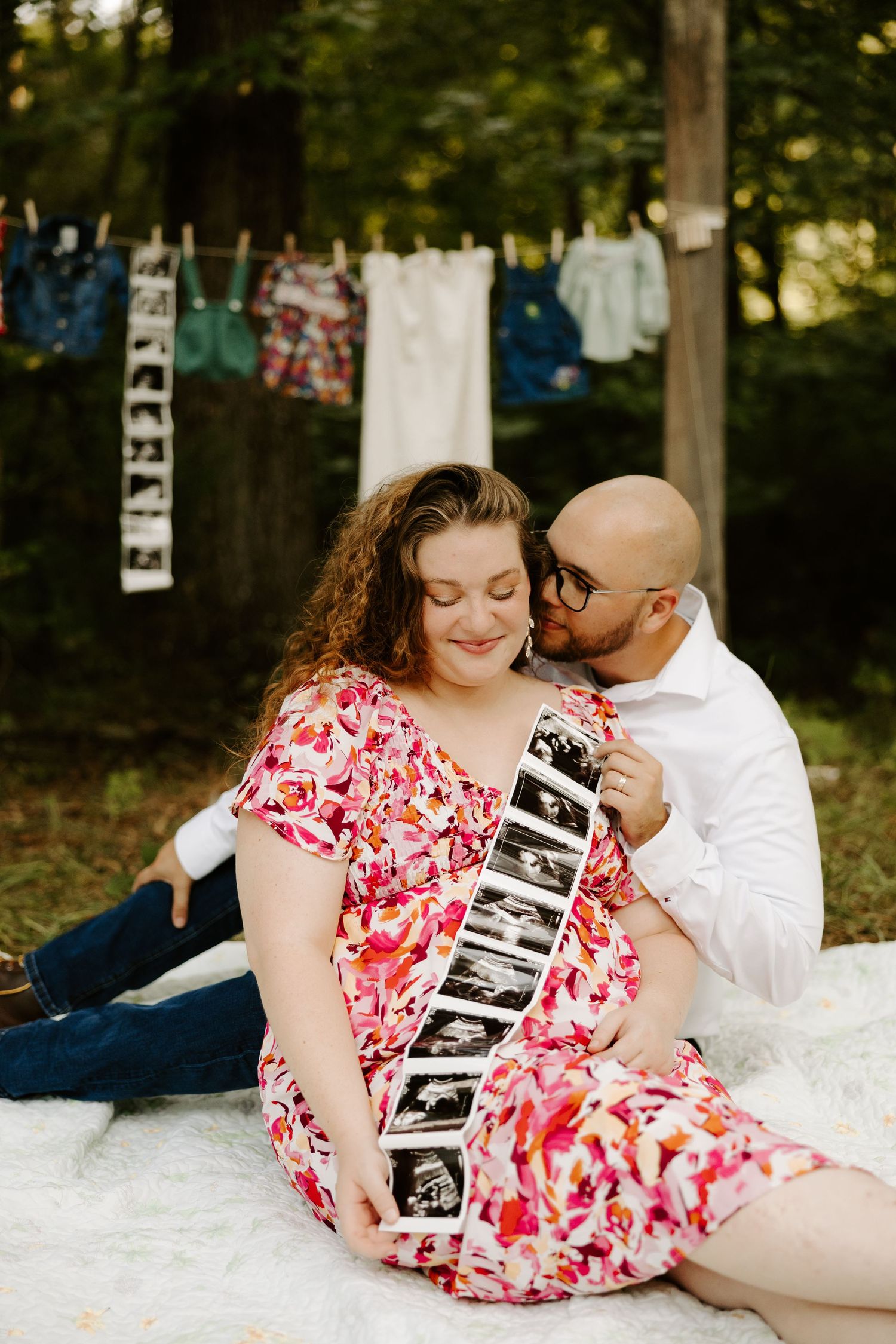 Pregnant couple sitting outdoors holding ultrasound photos with baby clothes hanging on a line behind them.