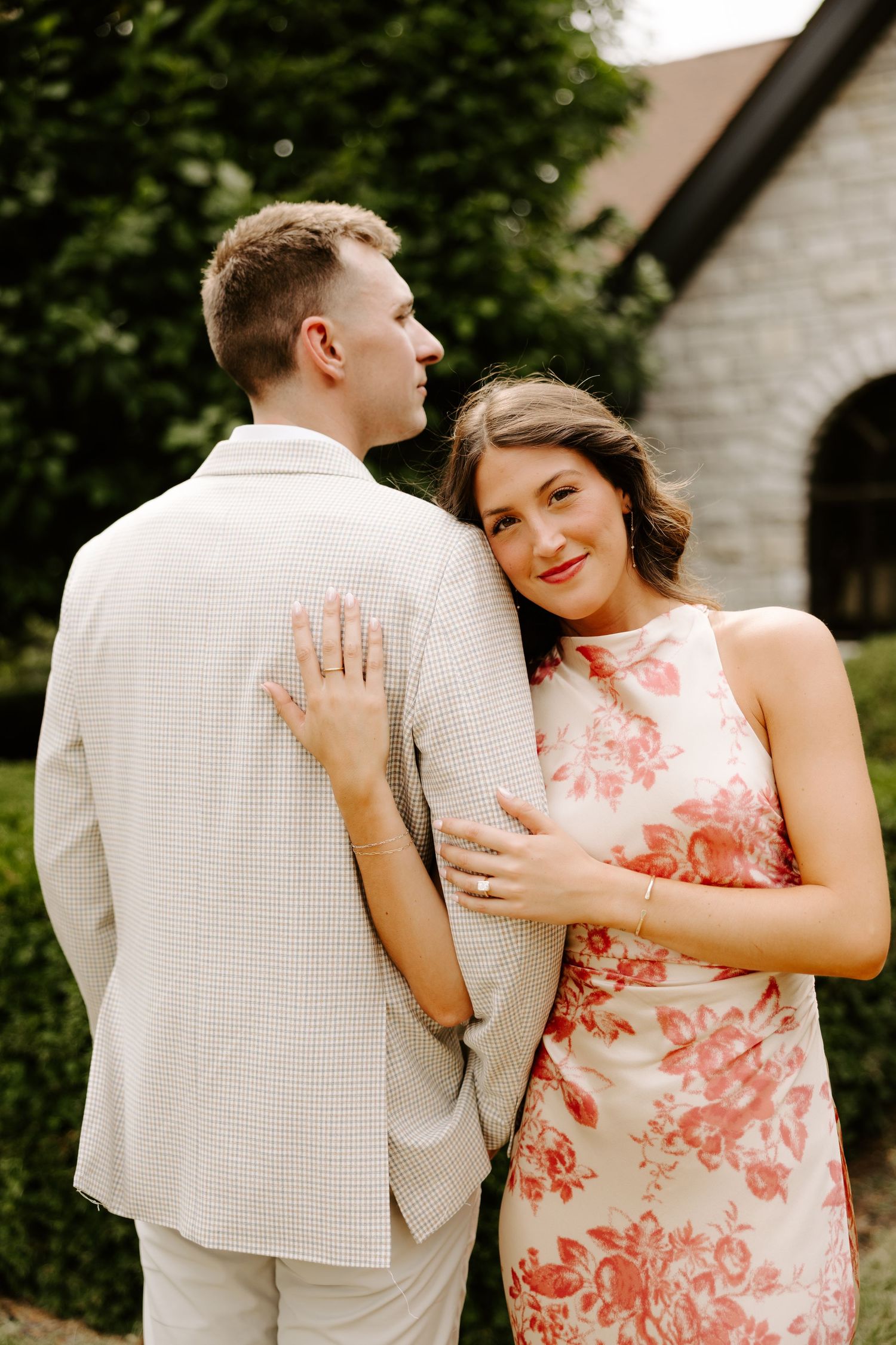 Couple embracing outdoors near stone chapel, woman in floral dress smiling over mans shoulder in cream suit.