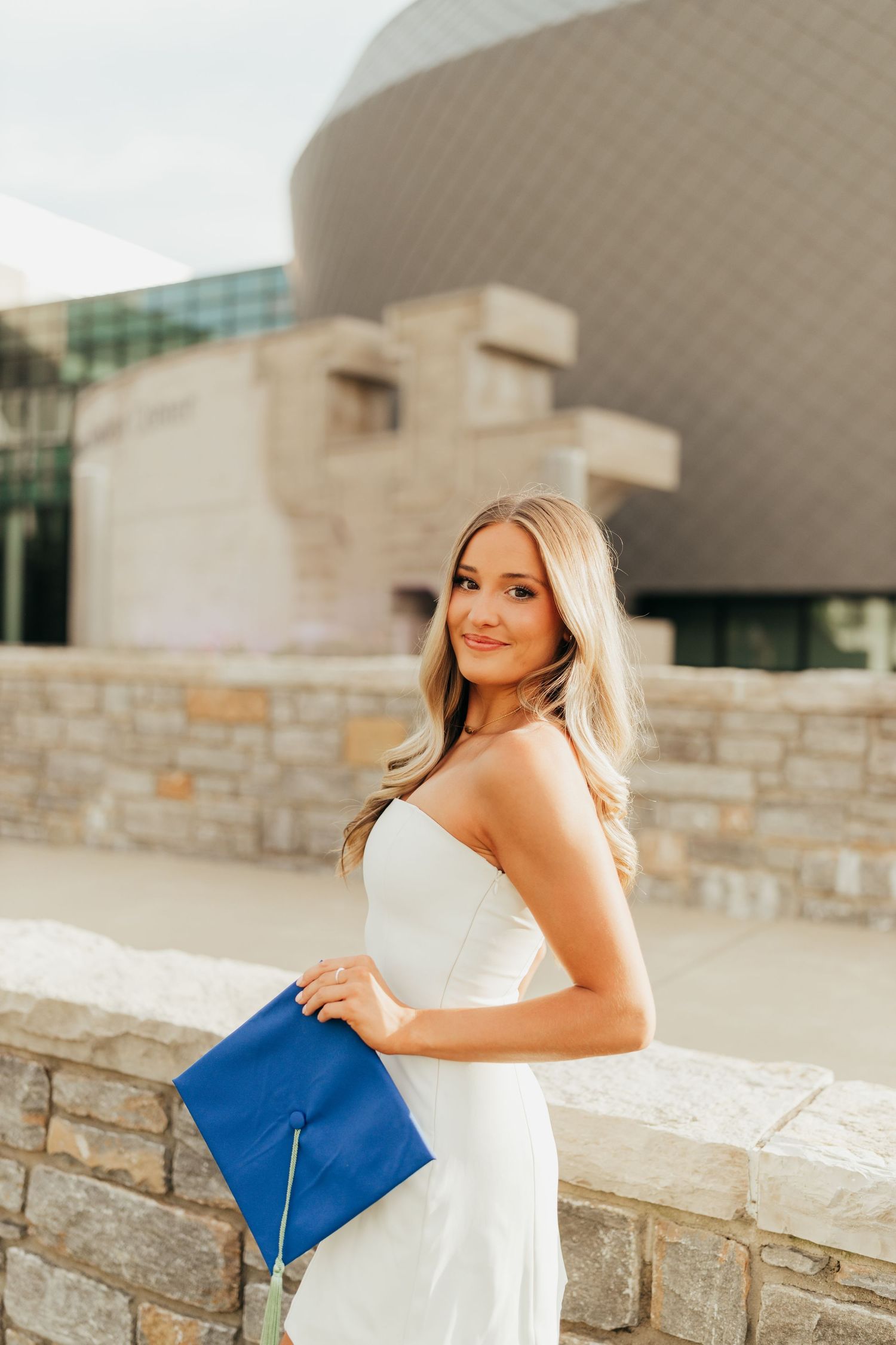 Graduate in strapless white dress poses with blue diploma by stone wall and modern architecture.