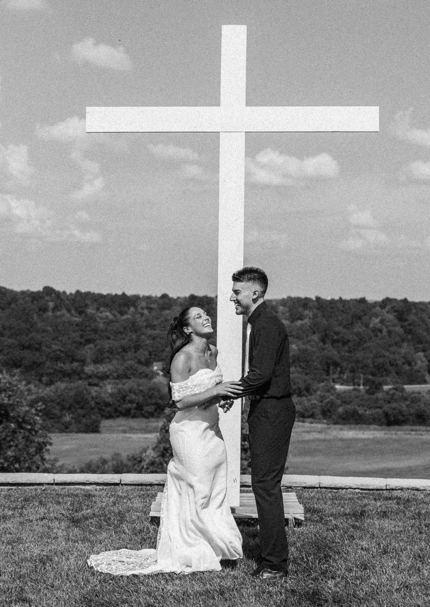 Black and white photo of a laughing bride and groom standing together beside a tall white cross outdoors.