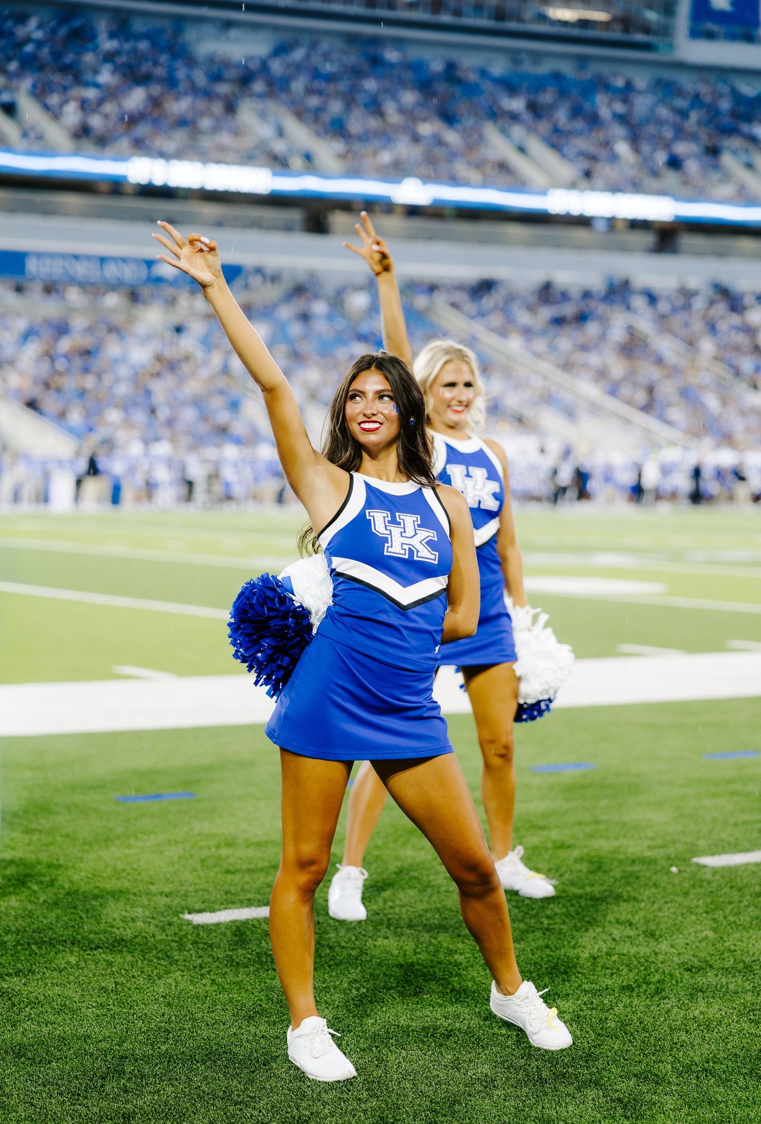 Two University of Kentucky cheerleaders perform on the sidelines with pom-poms during a Wildcats home football game.