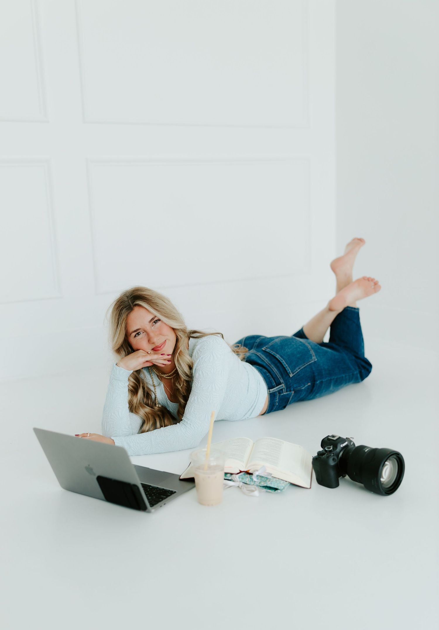 A smiling blonde woman in jeans and white top poses on a white floor with a laptop and camera.