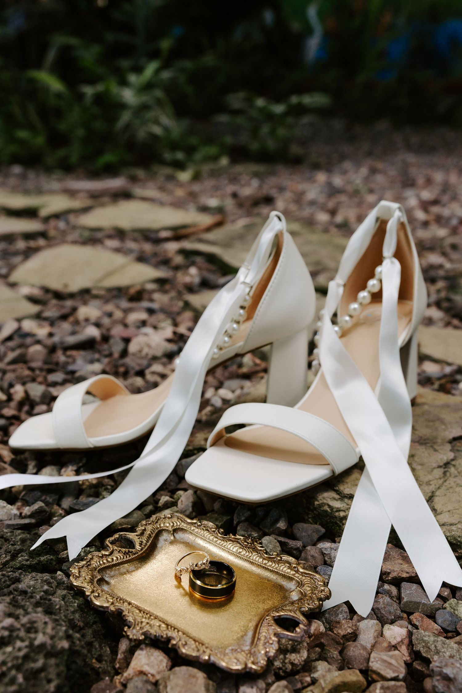 White bridal heels with ribbon ties and pearl details beside gold rings on decorative tray outdoors.
