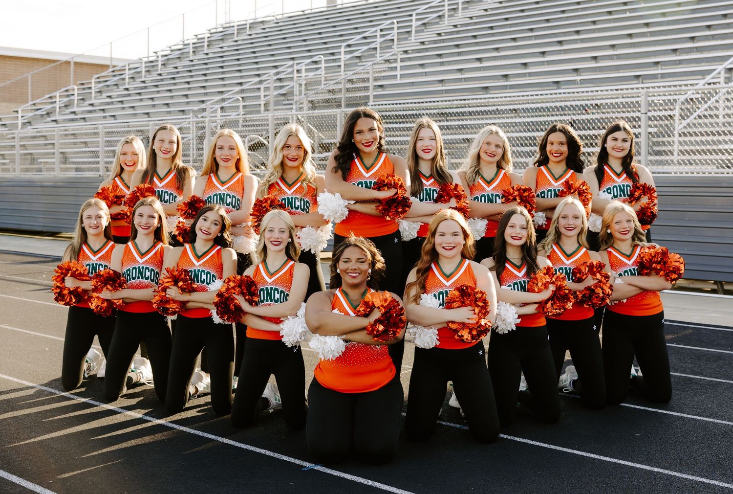 High school cheerleading squad in orange and black uniforms posing with pom-poms on stadium bleachers.