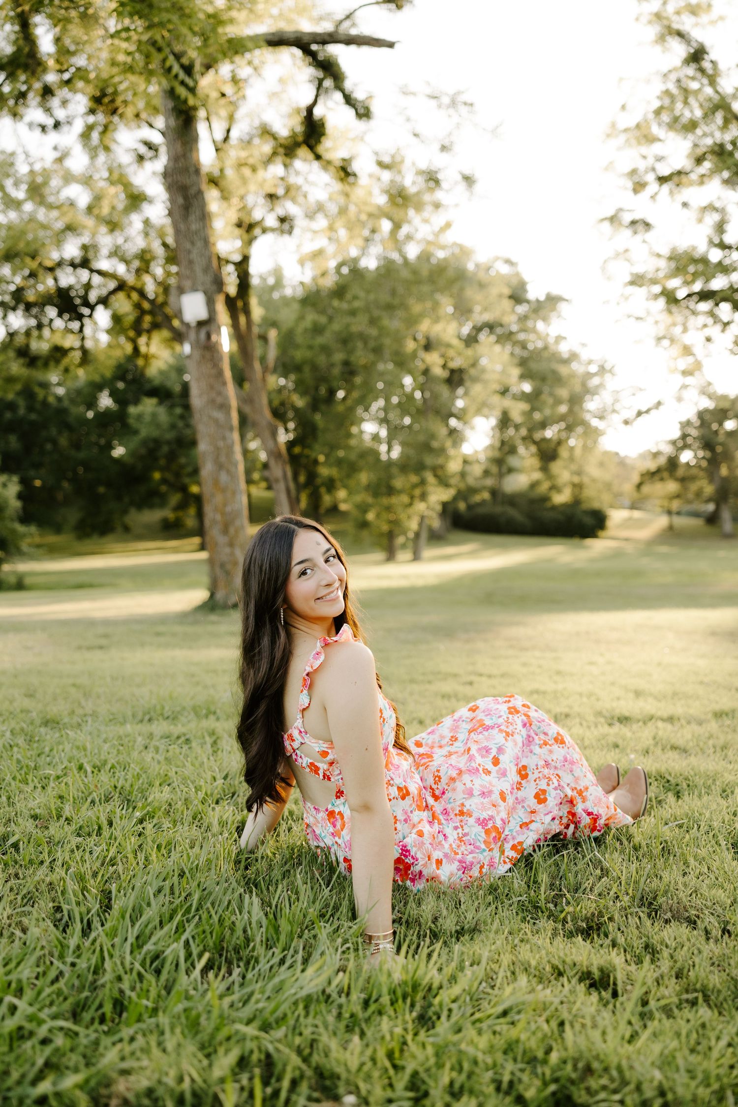 Smiling brunette girl in floral dress sitting on lush green grass in golden afternoon sunlight.