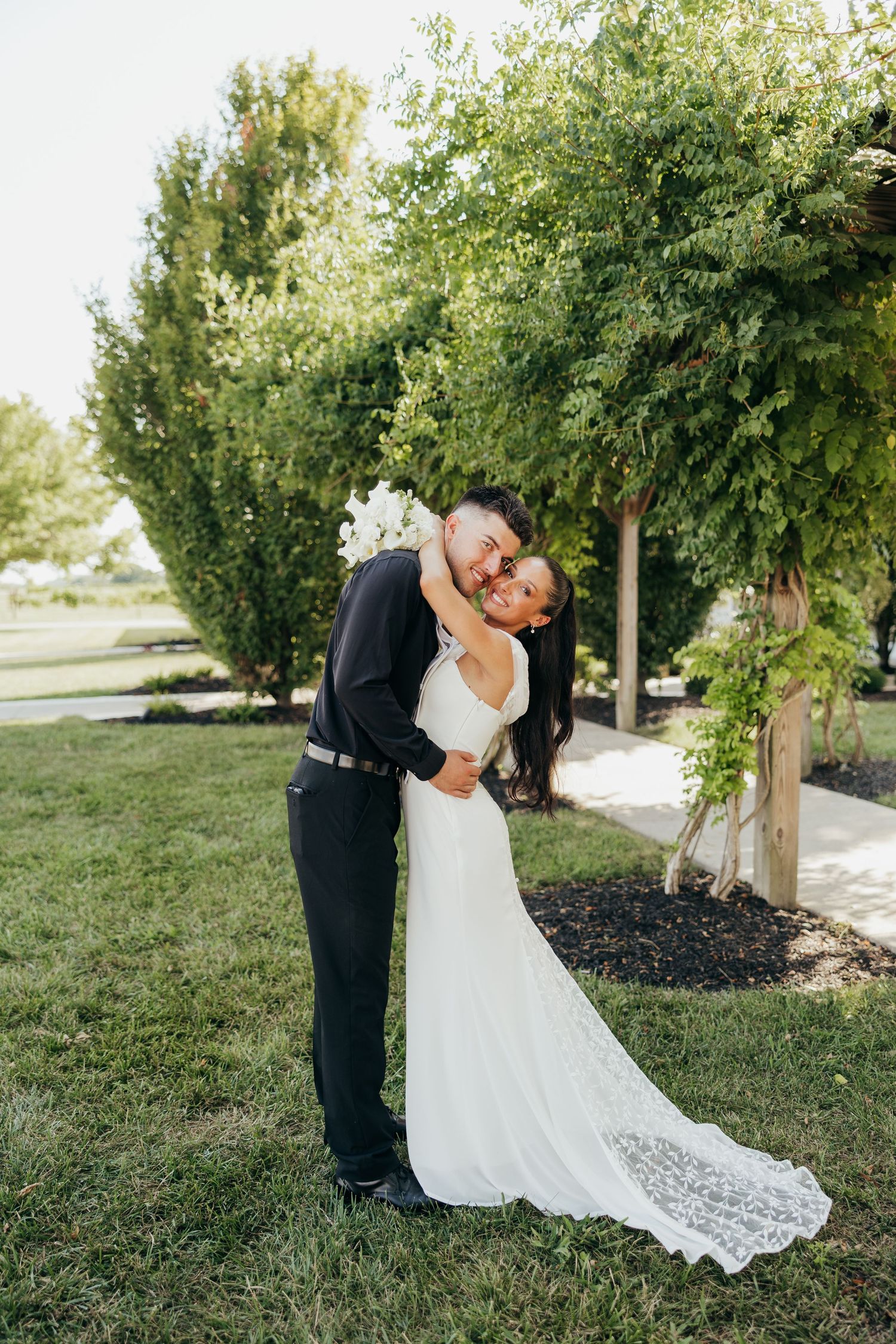 Groom embracing bride in elegant lace train wedding gown among green trees at outdoor venue.