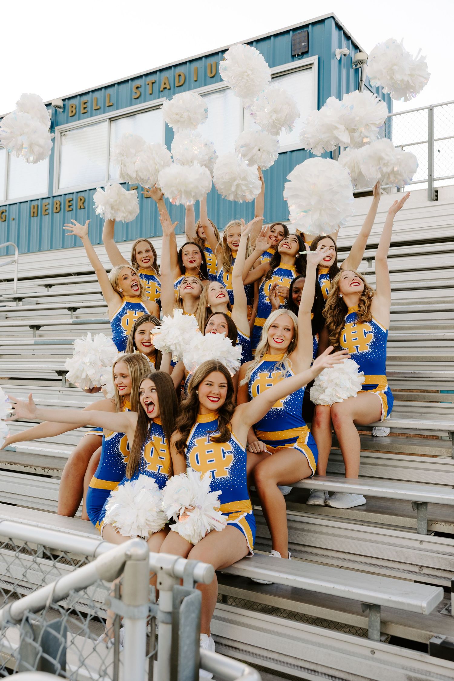 Henry Clay cheerleaders waving white pom-poms while posing on bleachers at a high school stadium.