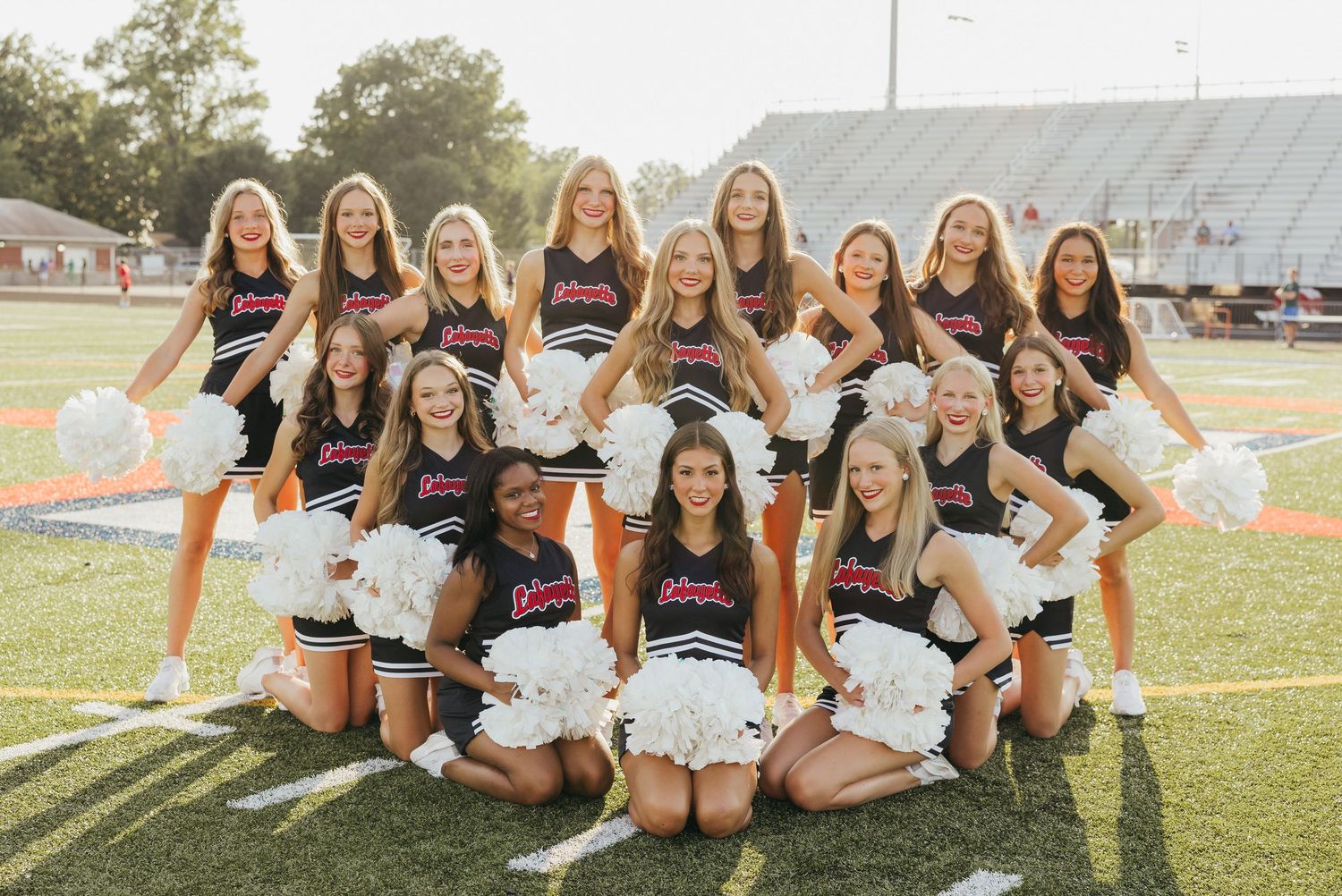 Cheerleading squad in black uniforms holding white pom-poms posing on a football field outdoors.